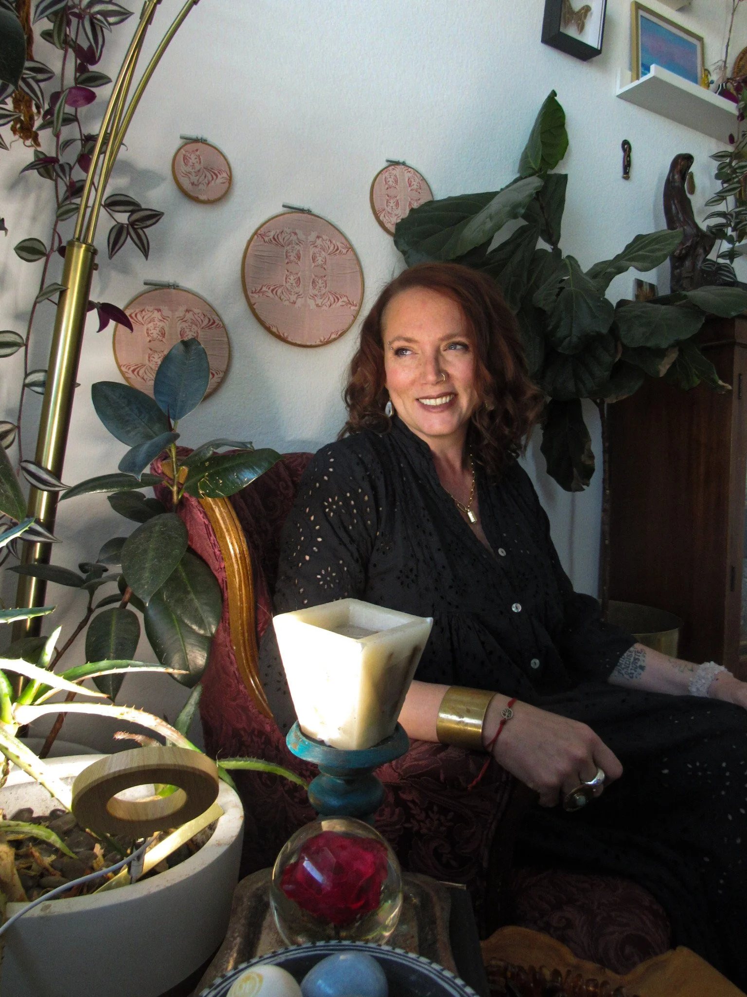 A woman with reddish curly hair and freckles sitting on a vintage red and gold upholstered chair, smiling and looking to her left in a cozy, decorated living room. She is wearing a black lace dress, a gold bracelet, and a ring. Behind her are green leafy plants and hanging embroidery hoops on the white wall. In front of her, there is a table with a candle holder, decorative objects, and potted plants.