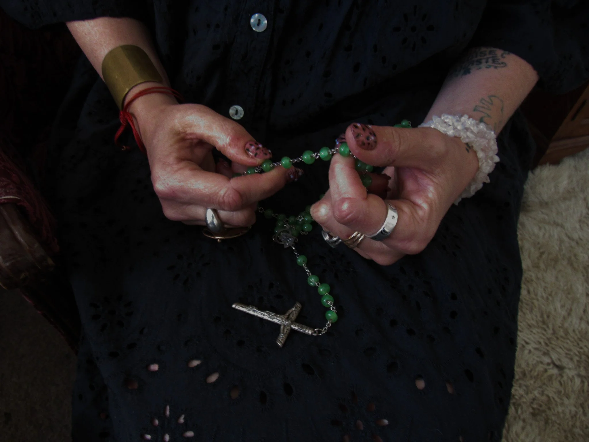 Hands holding a green beaded rosary with a silver crucifix, with tattoos, rings, a bracelet, and a white beaded wristband, against a black fabric background.
