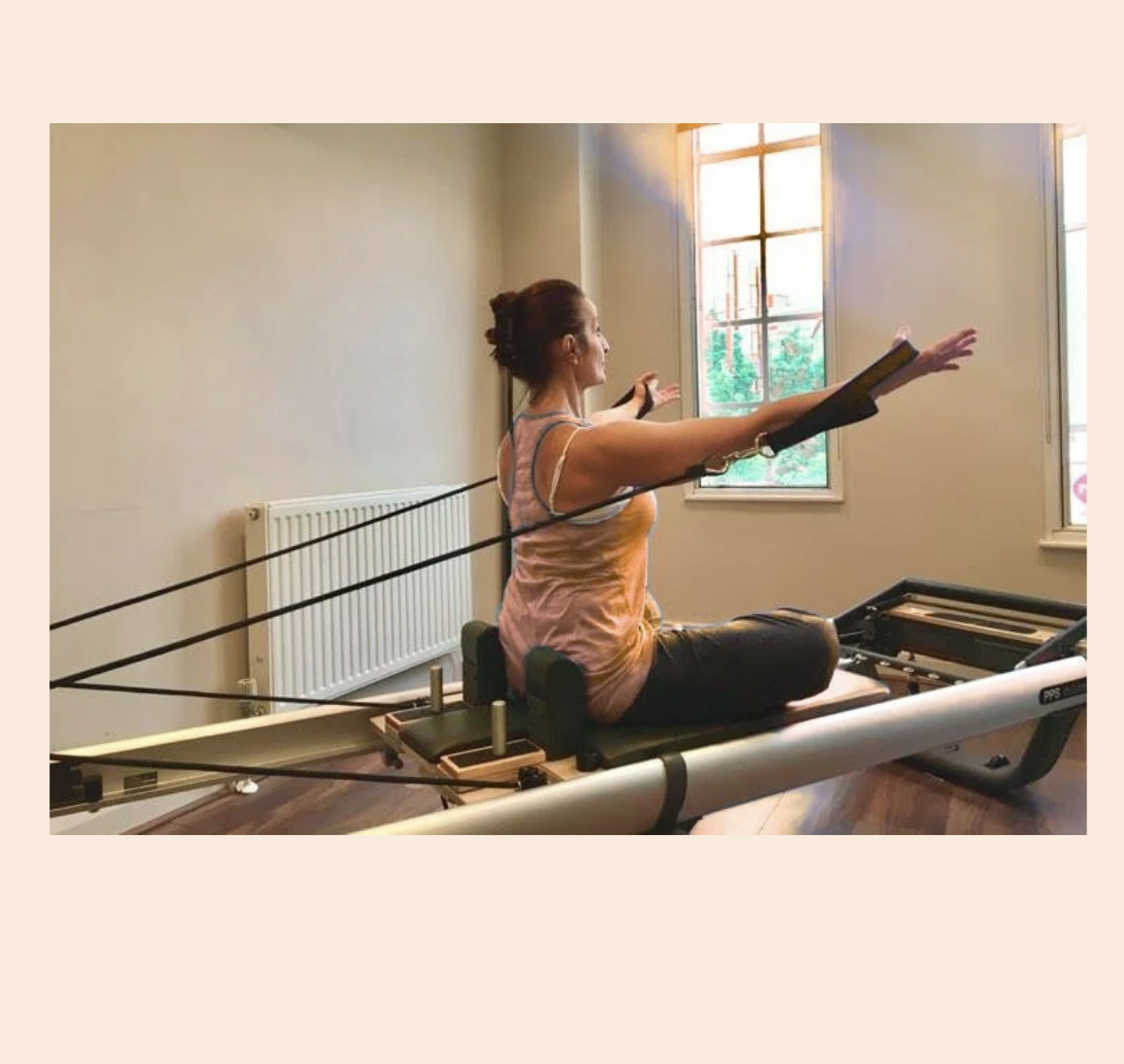 Woman practicing Pilates on reformer machine in bright room with large windows.