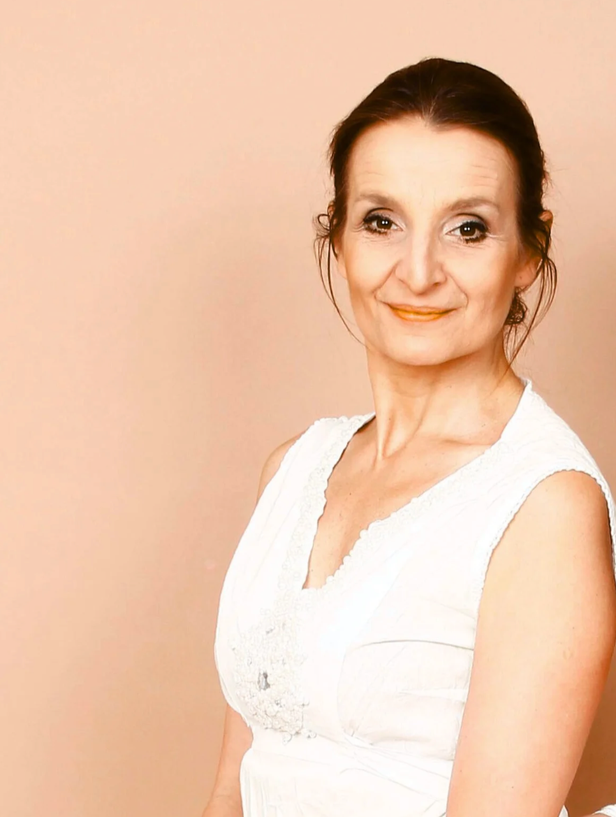 A woman with dark brown hair pulled back, wearing a sleeveless white top, smiling softly against a light peach-colored background.