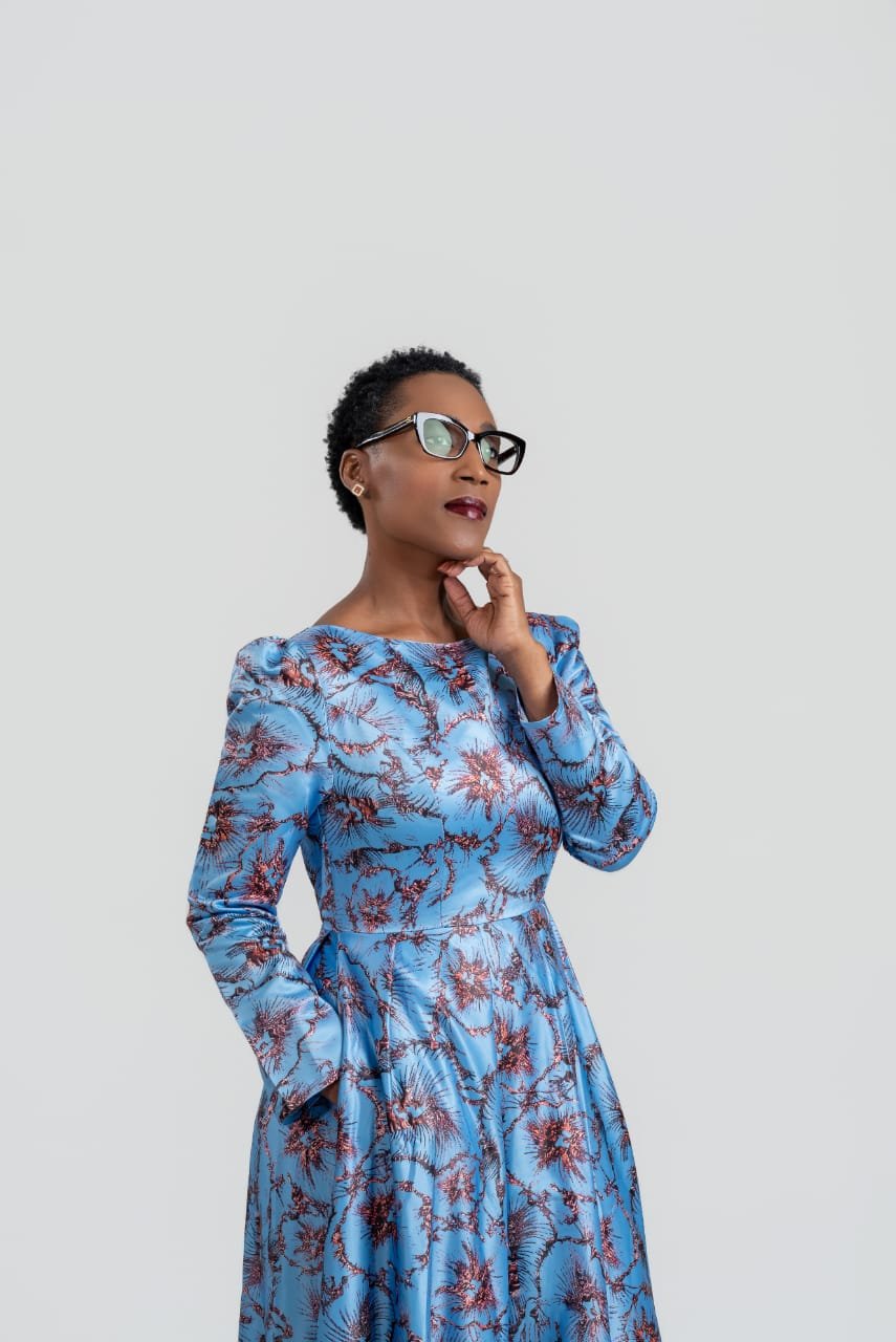 Elegant woman wearing a blue floral dress and glasses, posing thoughtfully against a plain light gray background.