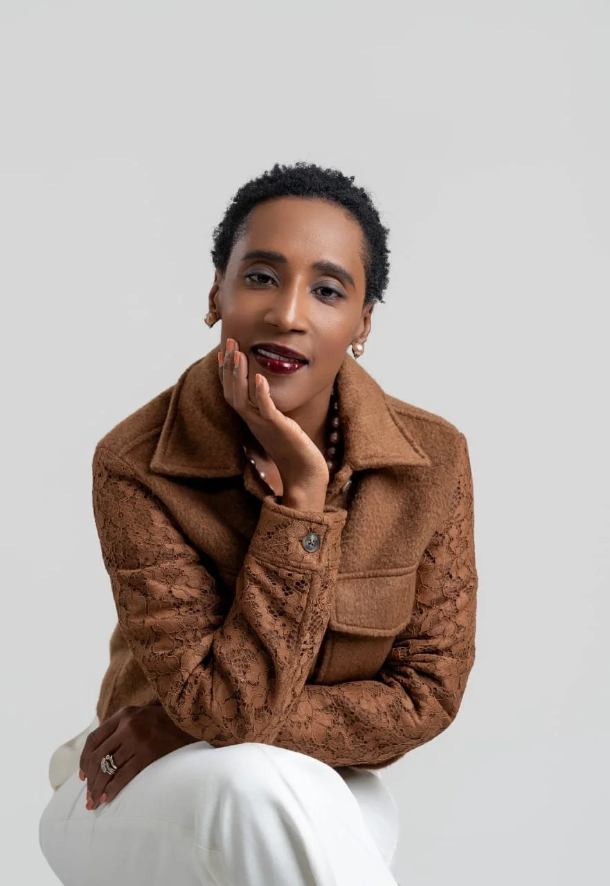 Portrait of a woman with short curly hair wearing a brown jacket with lace sleeves, white pants, and jewelry, posing against a plain light gray background.