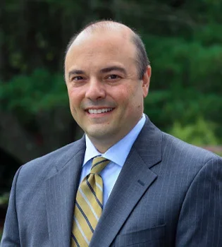 A middle-aged man in a gray suit, blue shirt, and yellow striped tie standing outdoors with greenery in the background.