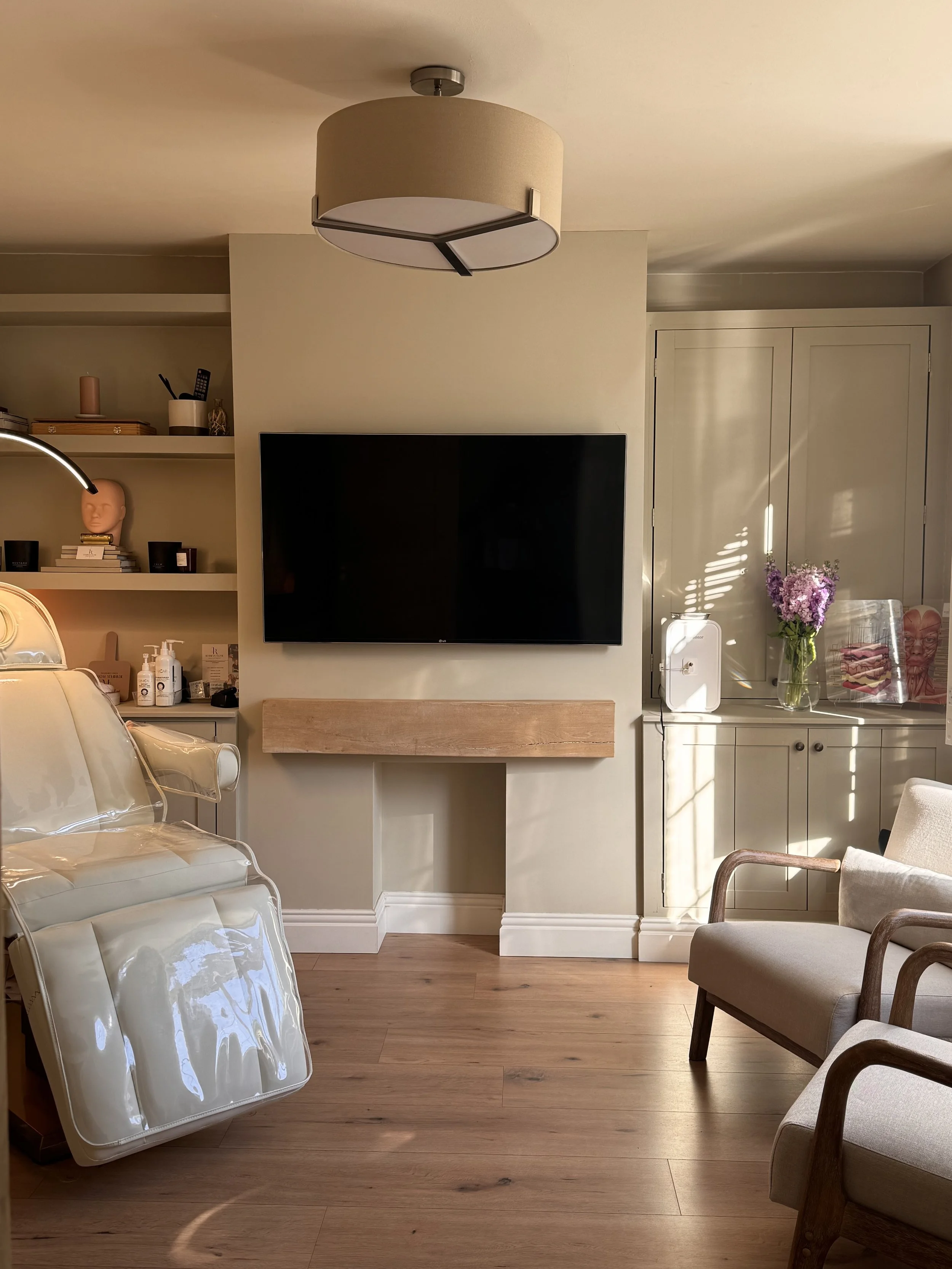 A living room with a wall-mounted flat-screen TV, a beige armchair, a wooden floor, and a cabinet with flowers and artwork. There are chairs with transparent covers, a shelf with decorative objects, and a ceiling light.