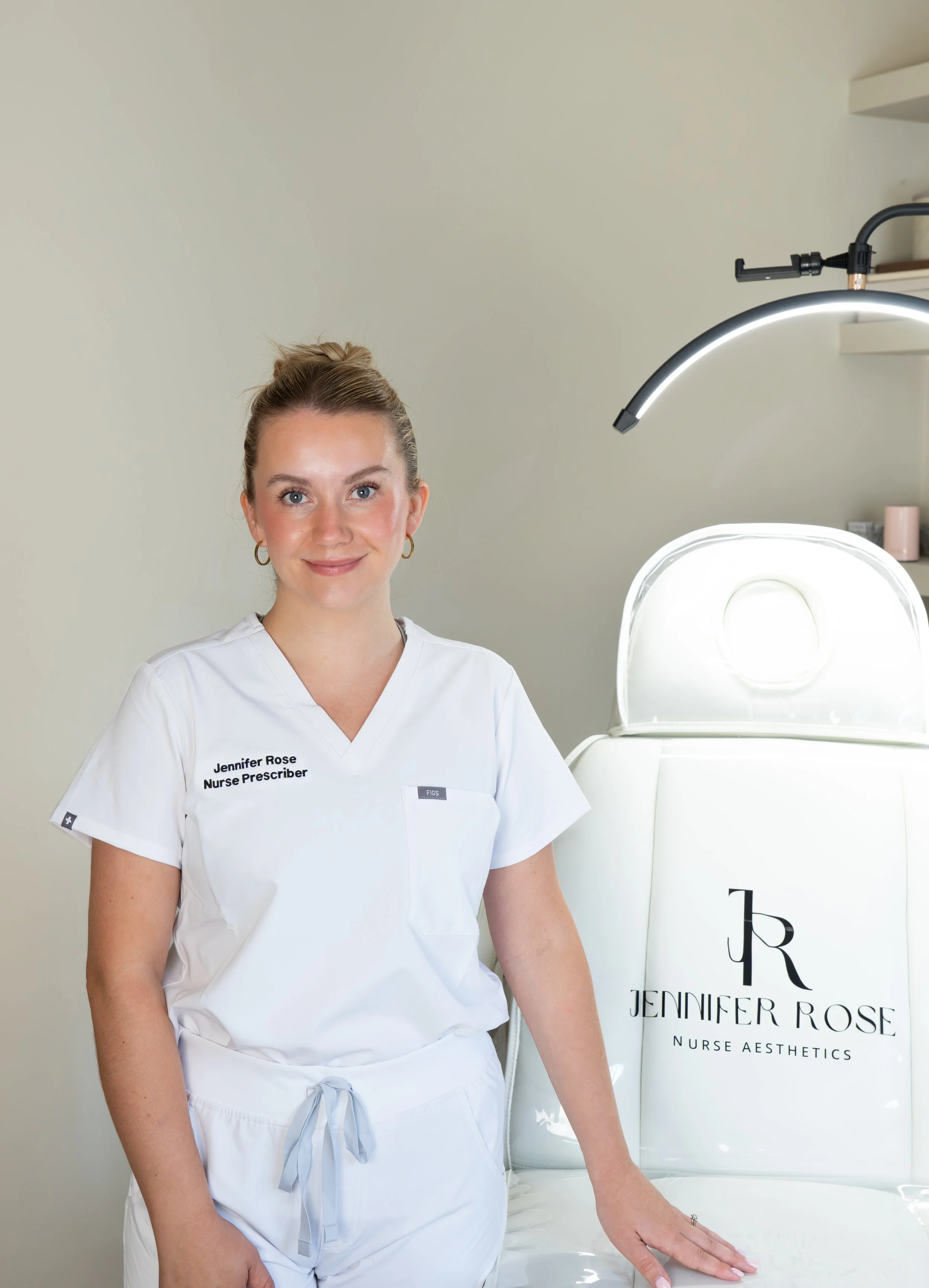 Professional woman in white scrubs with her name and title embroidered, standing next to a branded sign for 'Jennifer Rose Nurse Aesthetics' in a medical or spa setting.