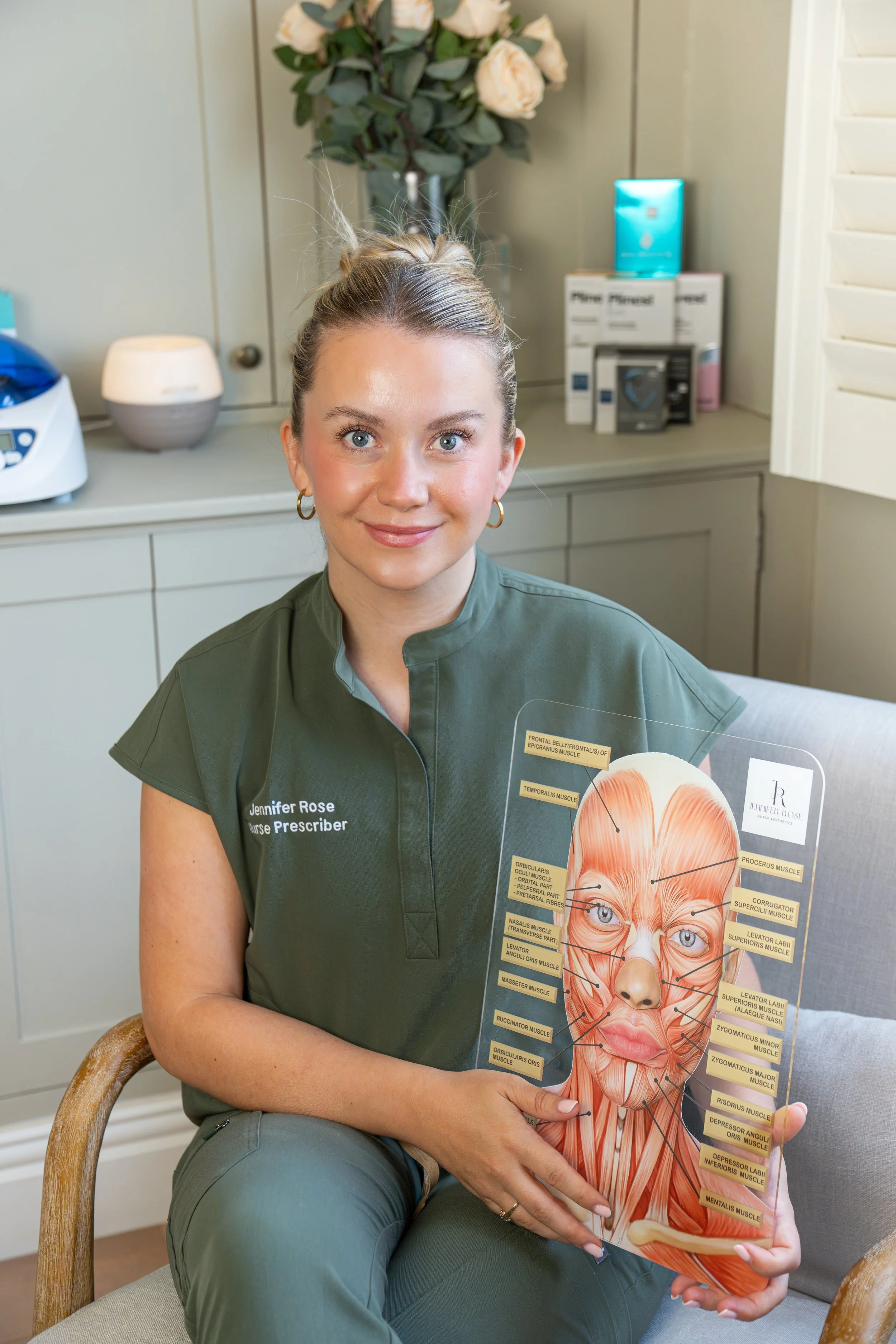A woman with blonde hair tied back, wearing a green medical uniform with name tag "Jennifer Rose" and gold hoop earrings, sitting on a beige and wooden chair, holding a detailed diagram of facial muscles. She is smiling slightly and looking directly at the camera. In the background, there is a small table with a vase of white roses, a box, and a device on top of a counter.