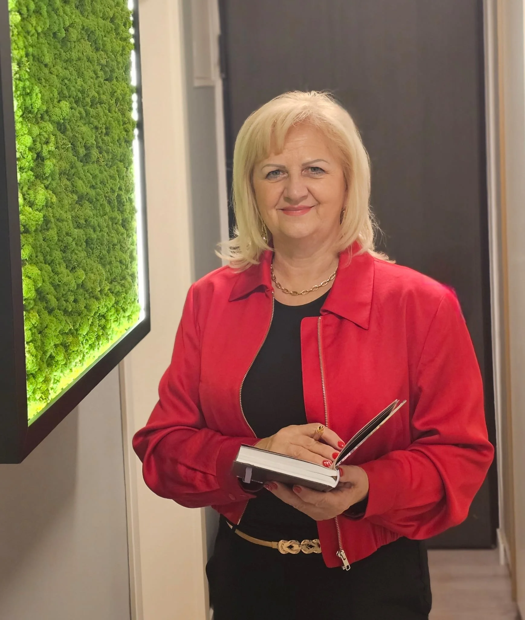 A woman with blonde hair wearing a red jacket and black top, holding a notebook, standing indoors near a frame with green moss or plant artwork.