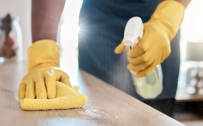 Person cleaning a wooden surface with a yellow cloth and spray bottle, wearing yellow gloves.
