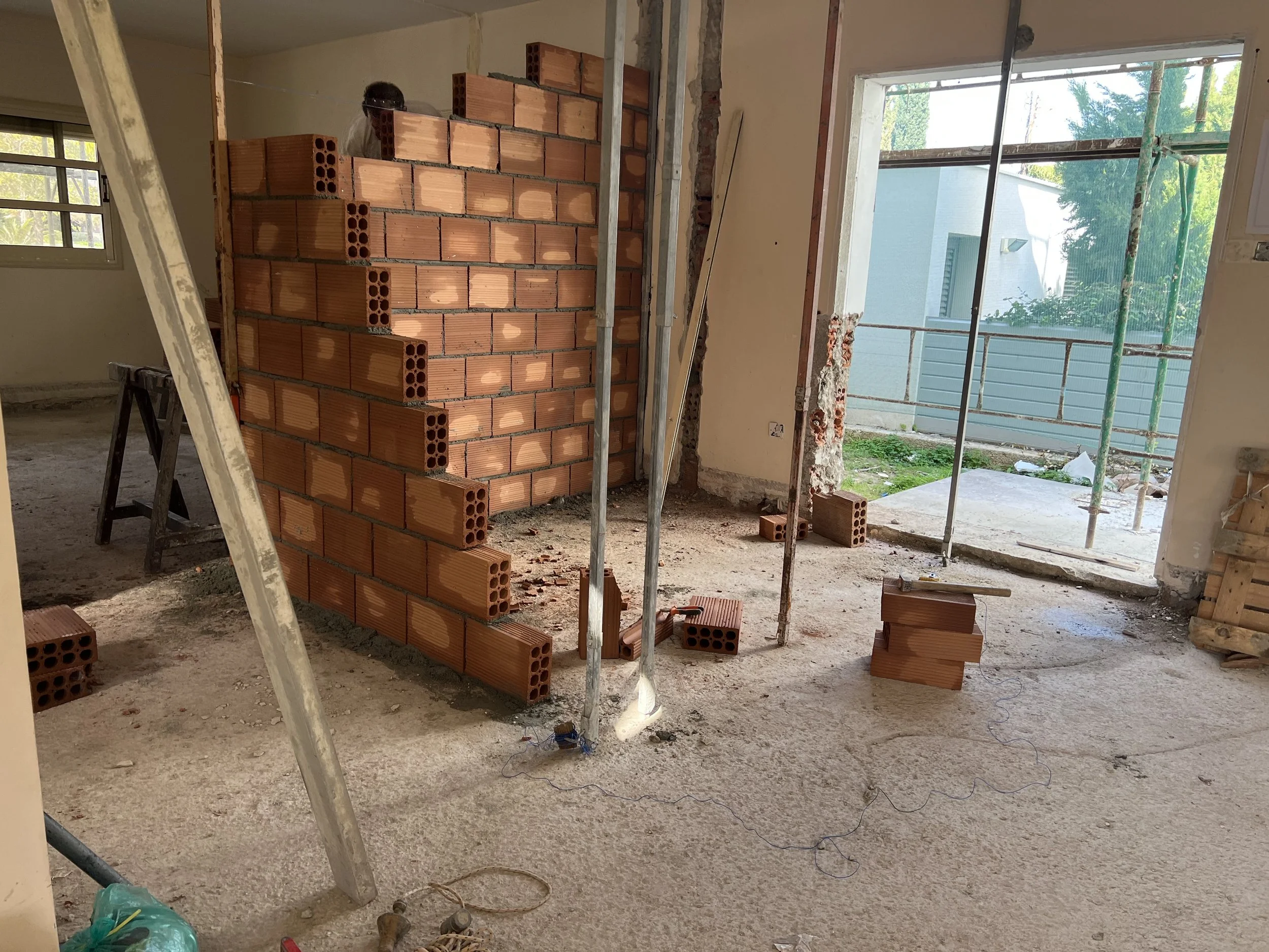 Interior of a house under construction with a brick wall being built, construction tools, and scaffolding near a sliding glass door opening to an outdoor area.