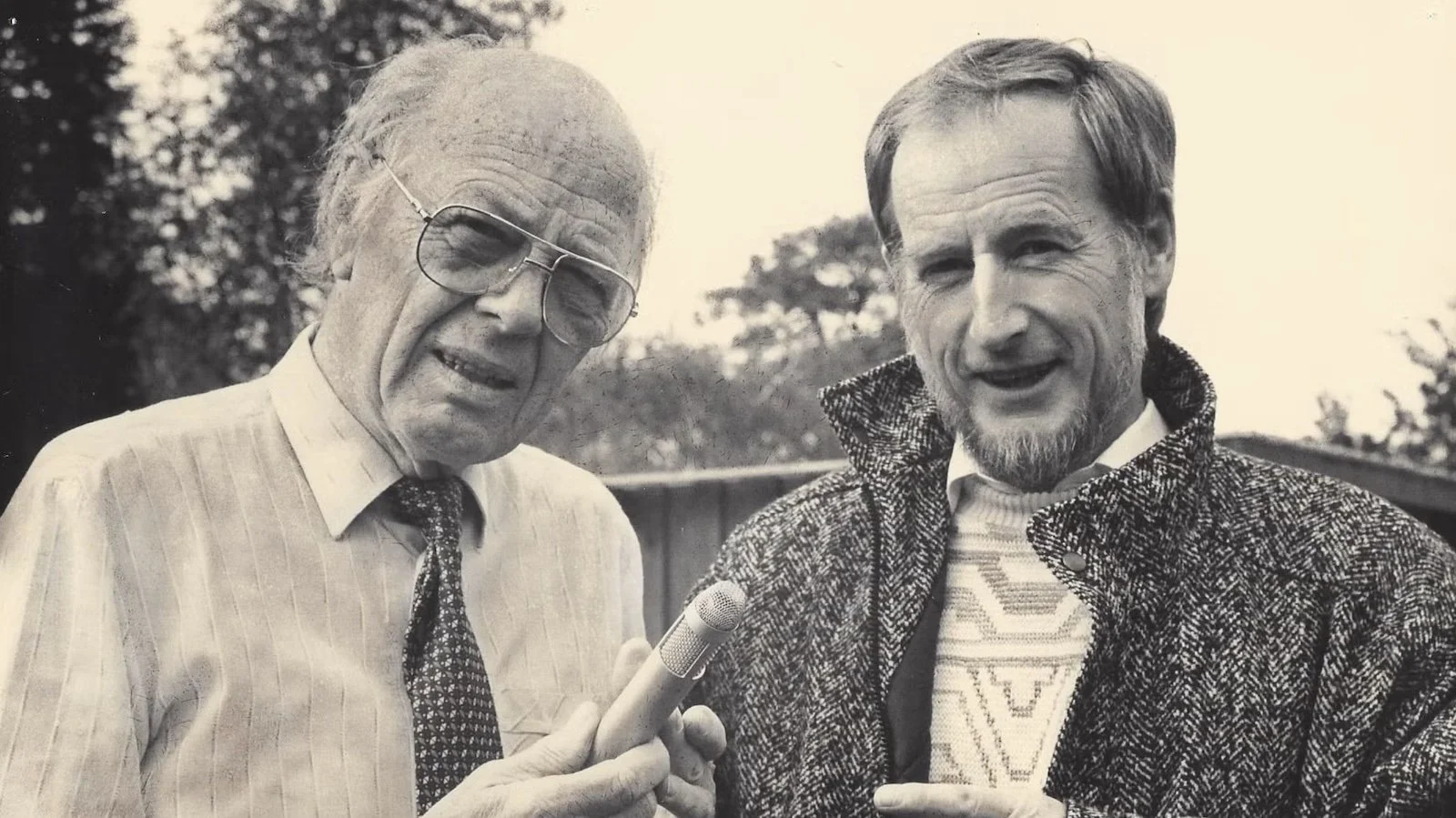 Black and white photo of two men outdoors, one holding a microphone, both smiling.