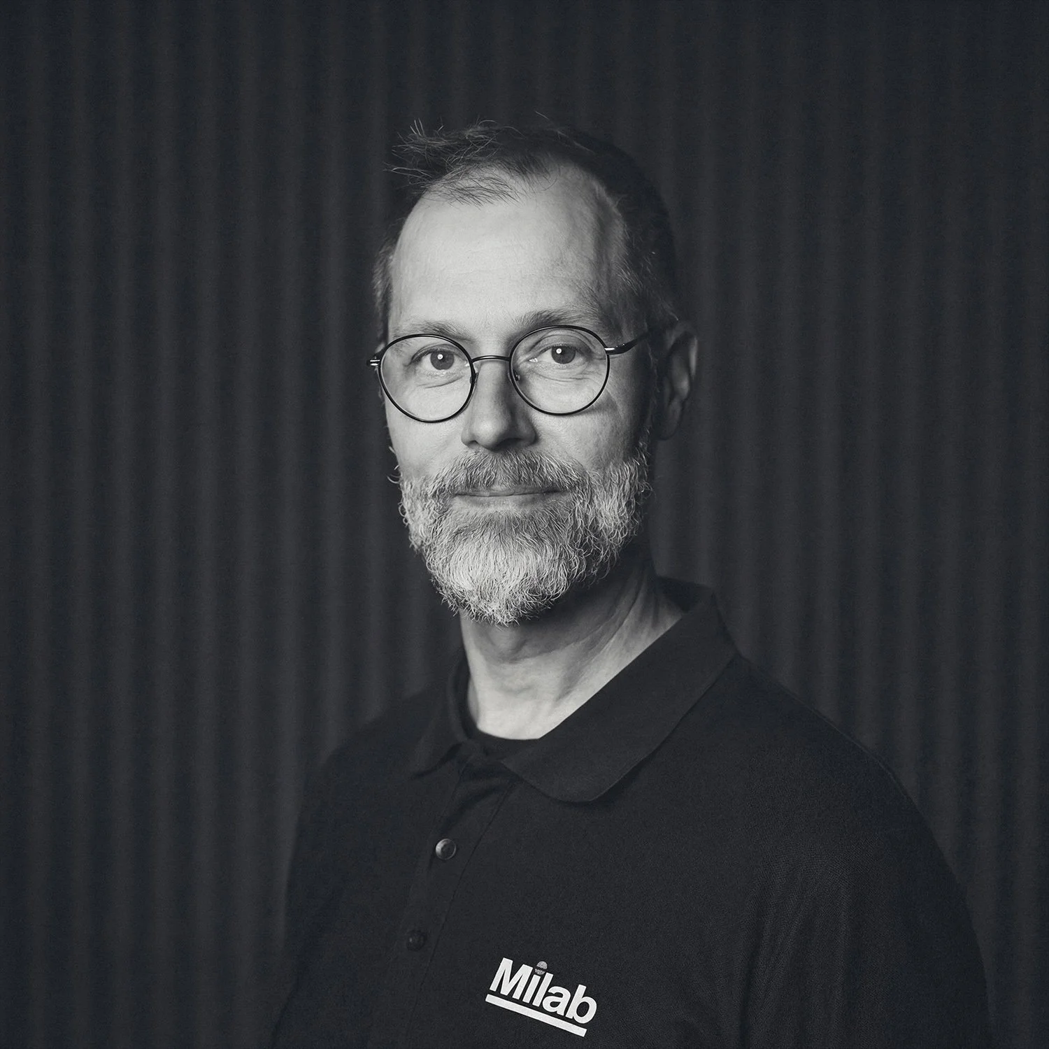 A black-and-white portrait of a middle-aged man with glasses, a beard, and short hair, wearing a black polo shirt with the logo 'Milab' on it, standing against a dark textured background.