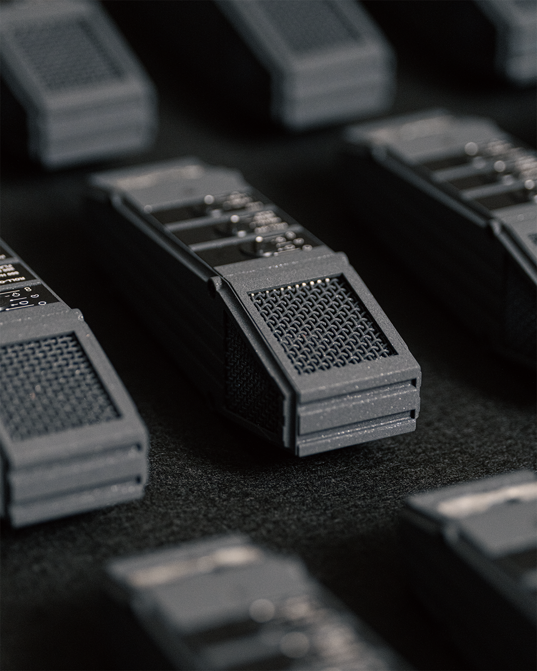 Close-up view of multiple black wireless microphone transmitters with textured grilles and control switches, arranged on a dark surface.