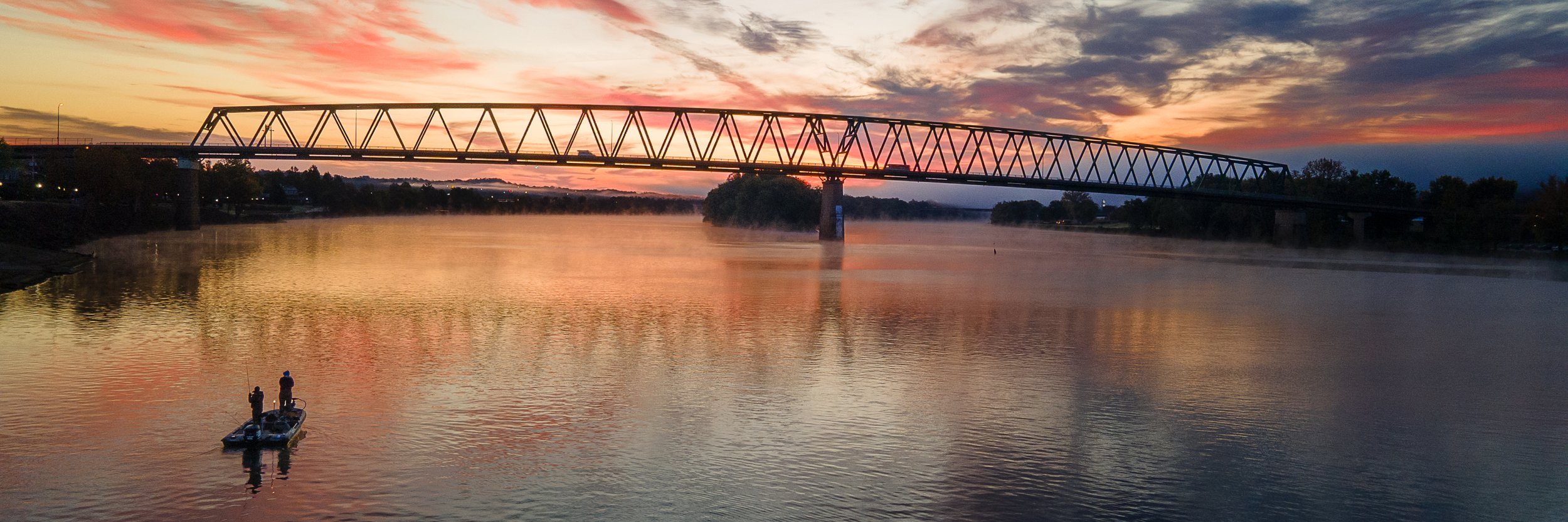 A sunset with a colorful sky over a river, a bridge stretching across the water, and two people fishing from a small boat.