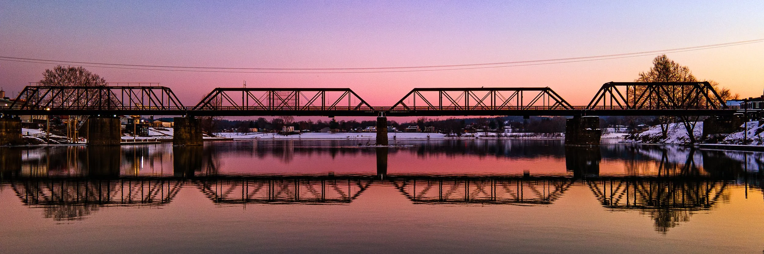 A metal truss bridge spanning over a calm river during a sunset or sunrise, with snow on the banks and trees in the background, and the bridge's reflection visible in the water.