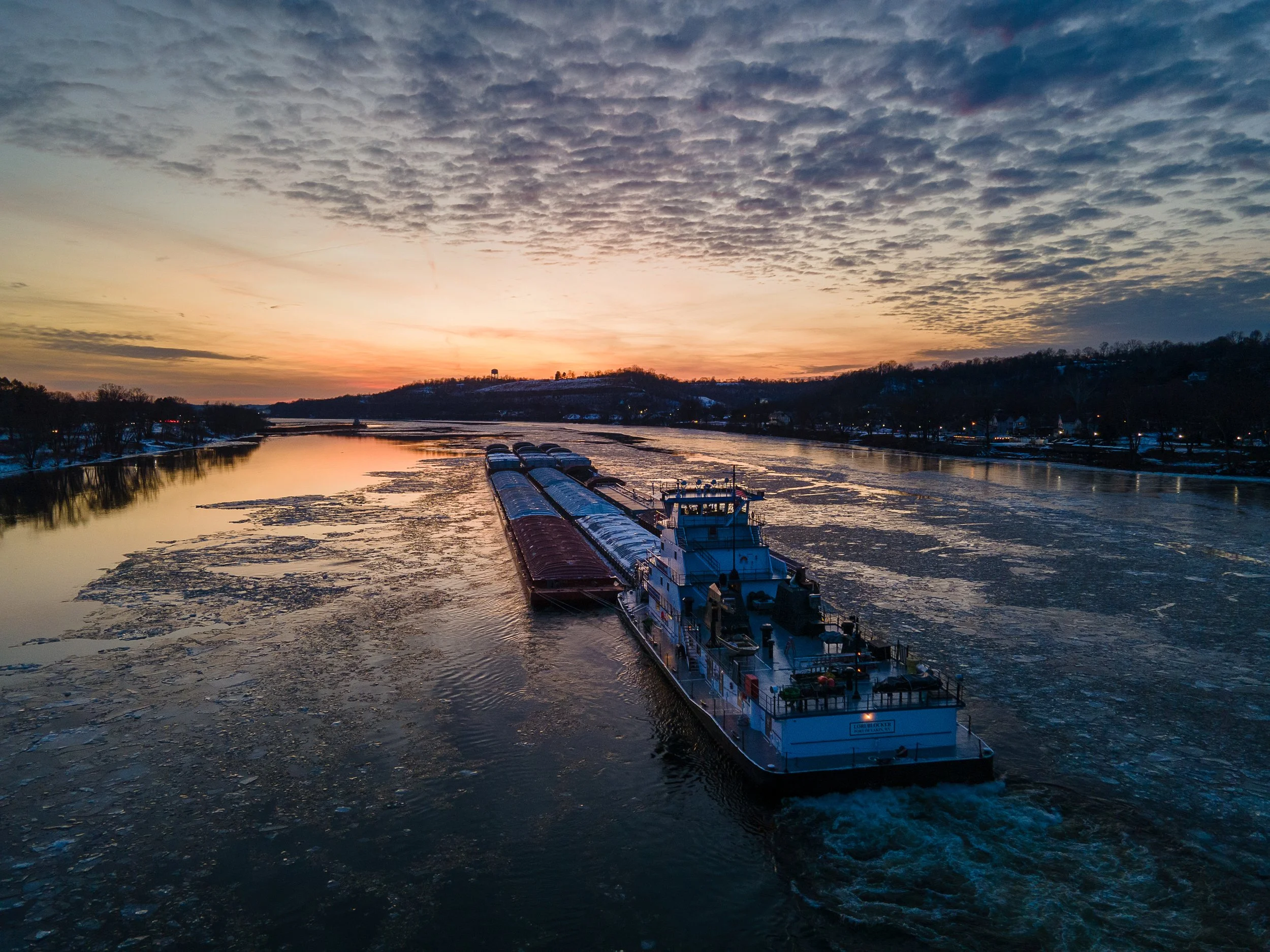 A large cargo ship sailing on a river during sunset with partially frozen water and a colorful sky.