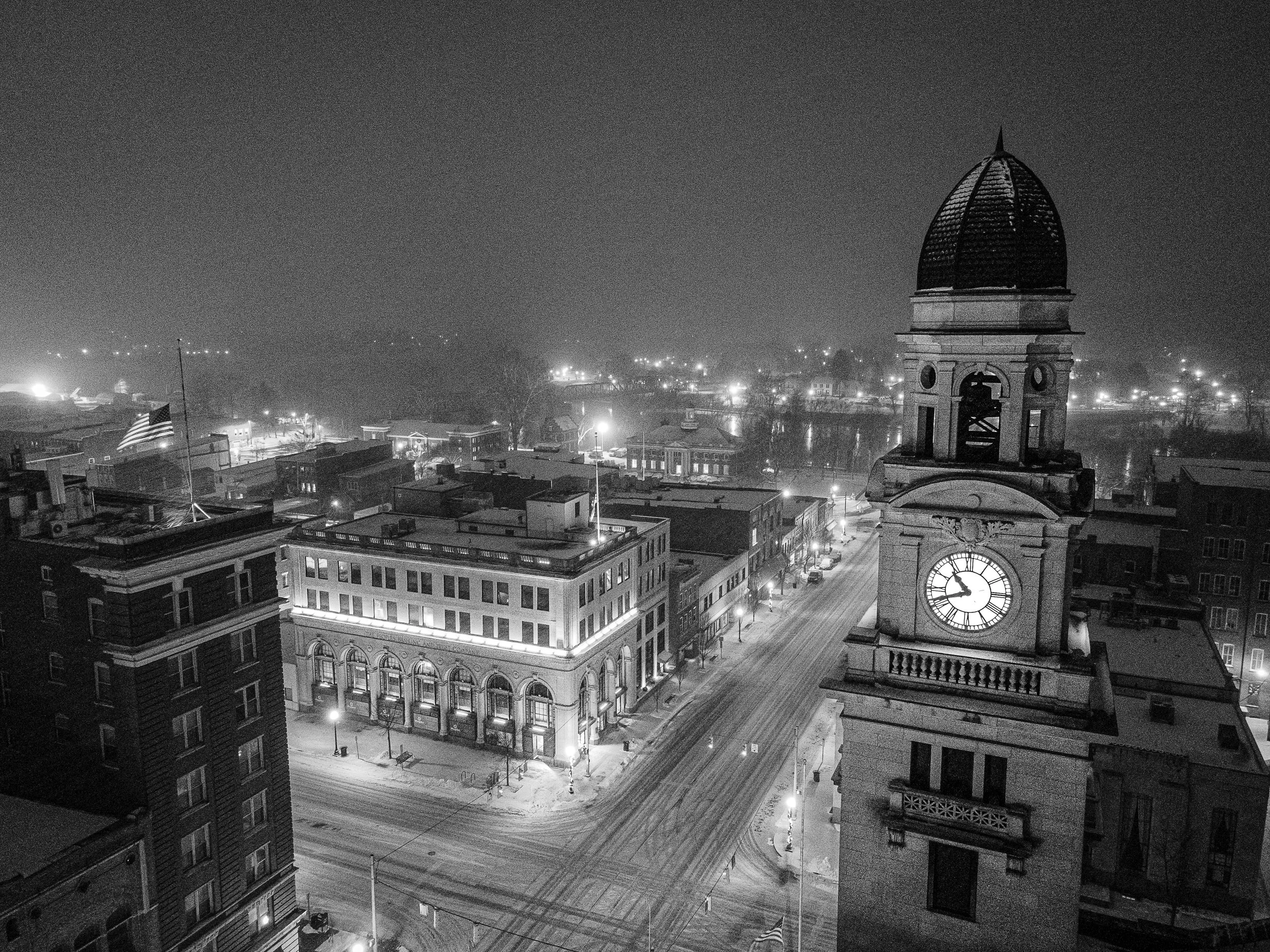 Nighttime view of a cityscape with a clock tower in foreground, showing 12:07, and buildings covered in snow, with streetlights illuminating the streets and a flag waving near the buildings.