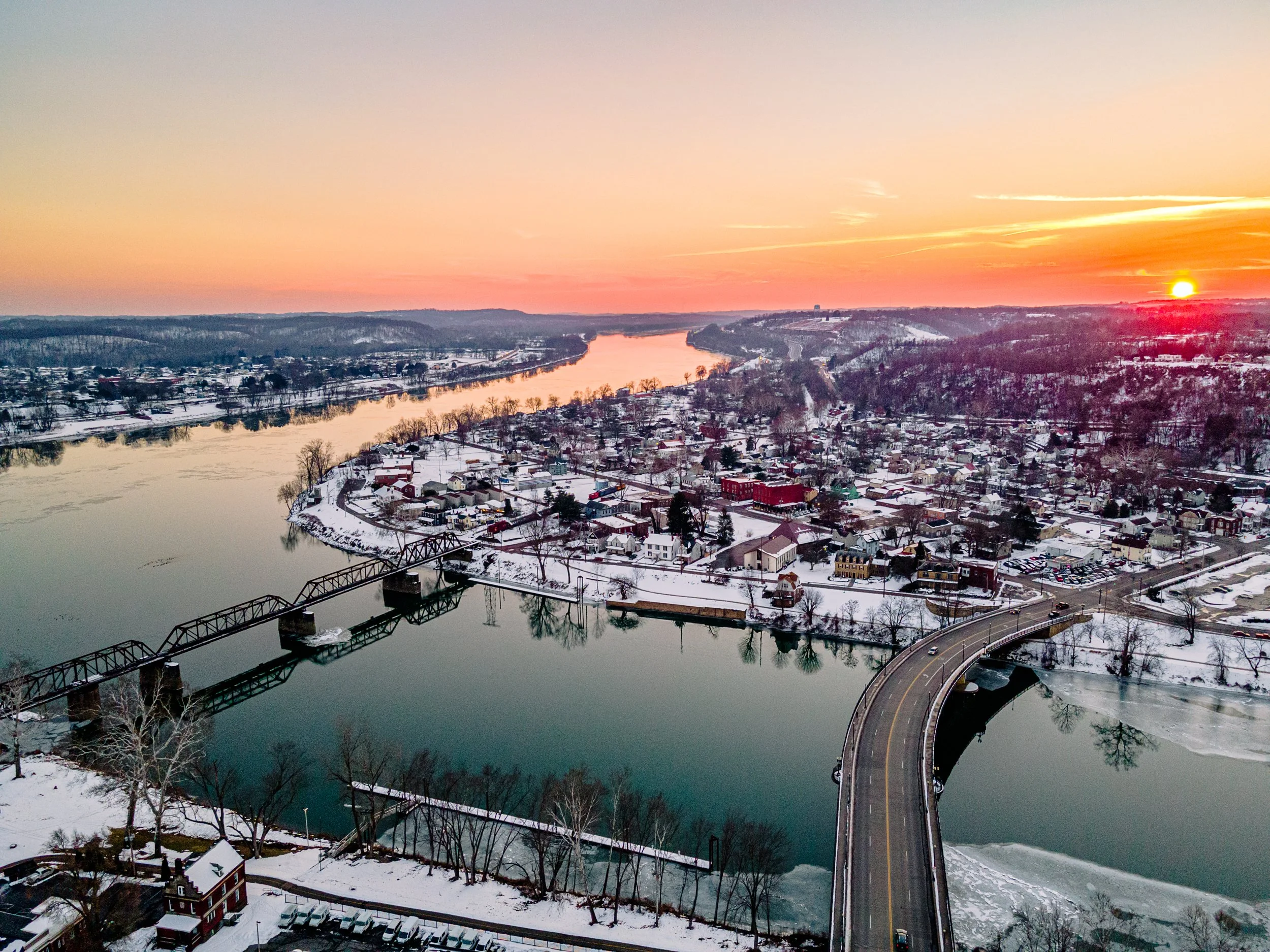 Aerial view of a snowy town at sunset with a river, bridge, and hillside houses.