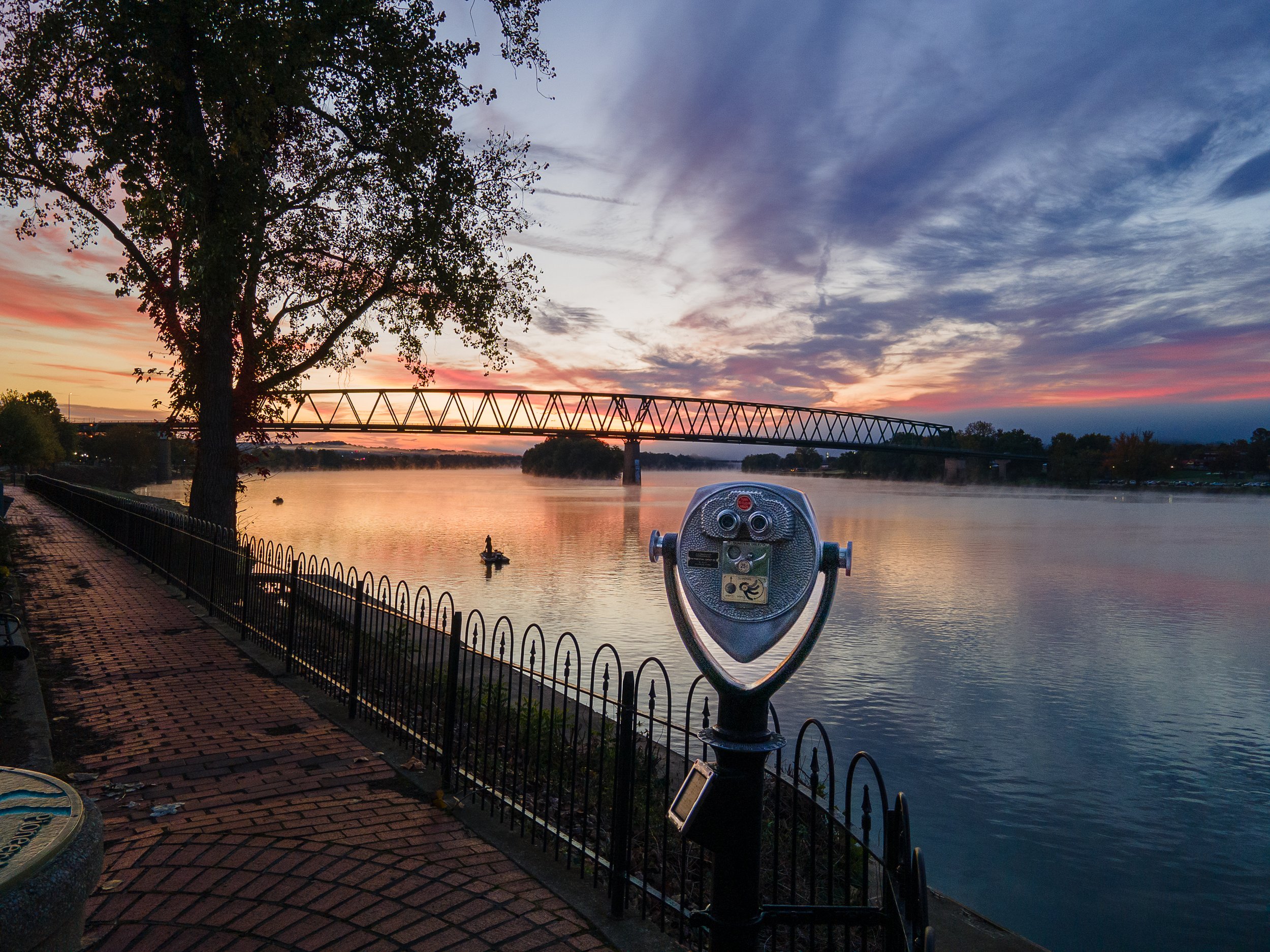 A river scene at sunset with a bridge in the background, a tree on the left, a paved walkway with a metal fence, and a coin-operated binocular viewer in the foreground.