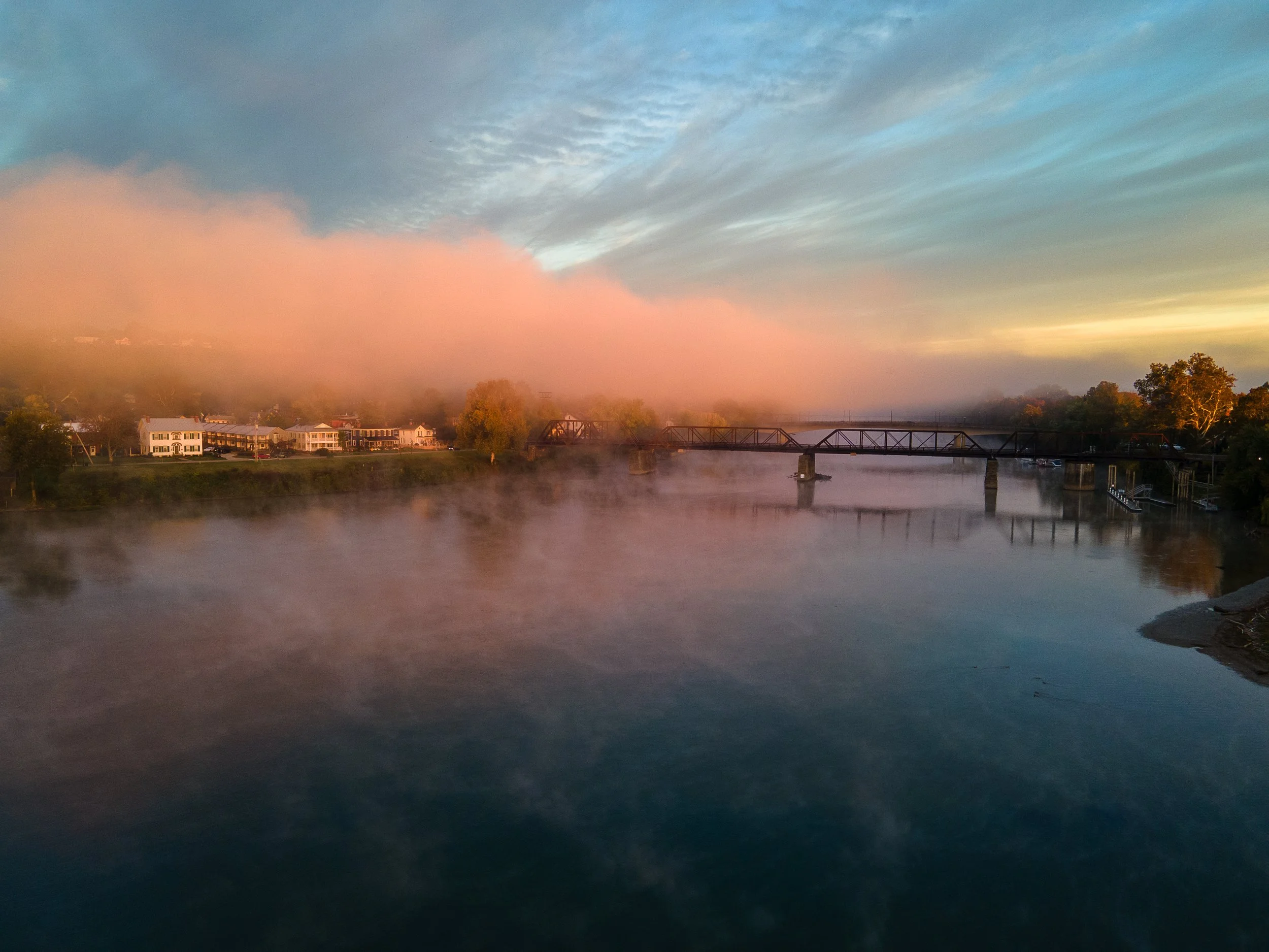 A river with mist rising from the water at sunrise, with houses on the left, a bridge crossing the river, and trees on the right, under a sky with scattered clouds.