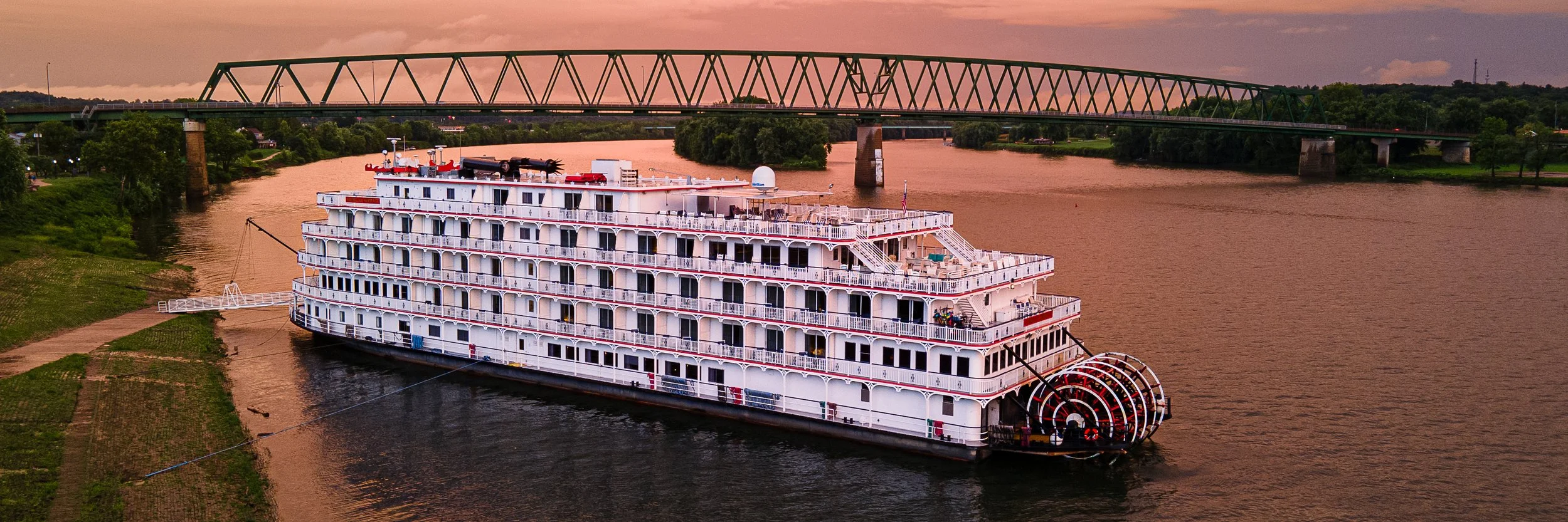 A large white river cruise boat with multiple decks docked on a wide river at sunset, with a highway bridge overhead and lush green trees in the background.