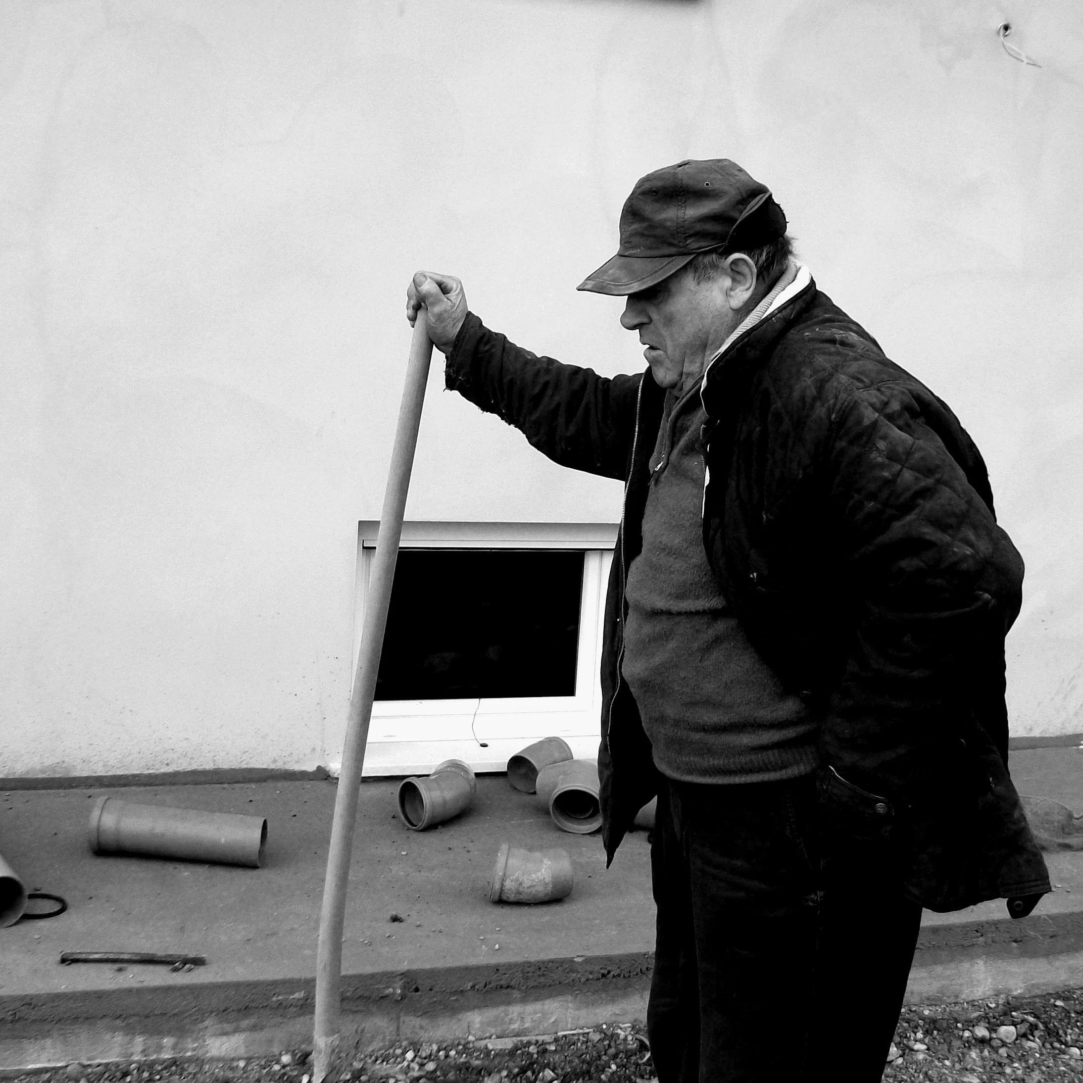 Uomo anziano con cappello e giacca scura posa con un'asta in una scena all'aperto, con tubi di plastica rotti e un'apertura di finestra sul muro dietro di lui.