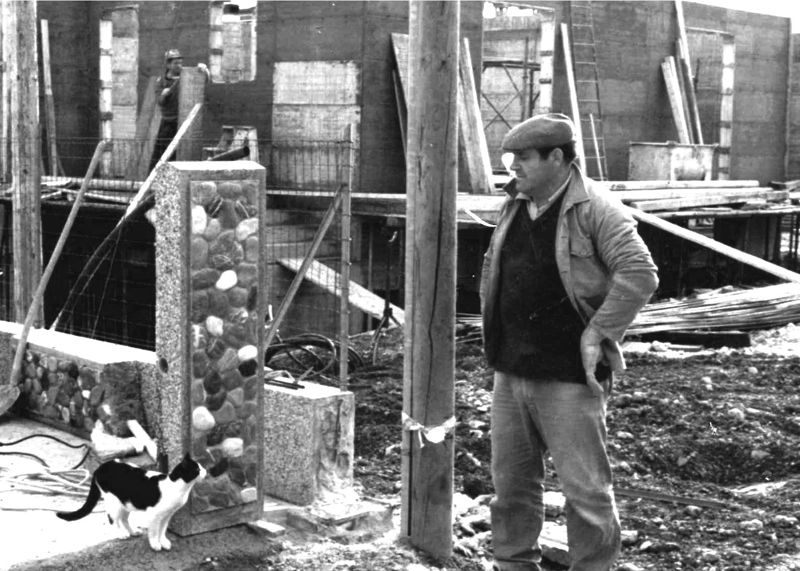 Uomo con cappello e giacca descriptiona un cane nero e bianco mentre osserva una parete di sassi colorati in un cantiere edile all'aperto, con recinzioni e strutture di legno sullo sfondo.