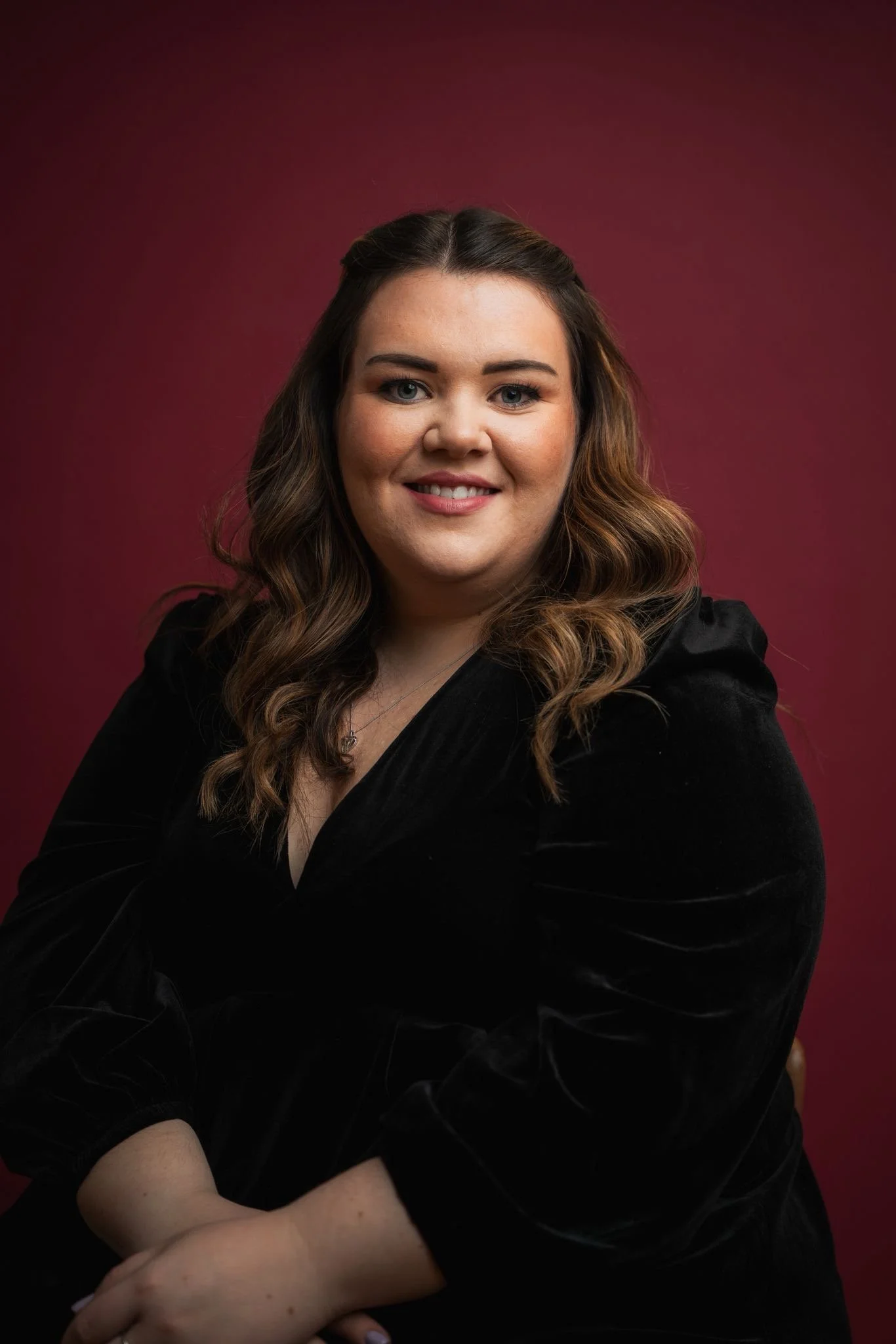A woman with long brown hair in loose curls, wearing a black top, smiling at the camera, with a maroon background.