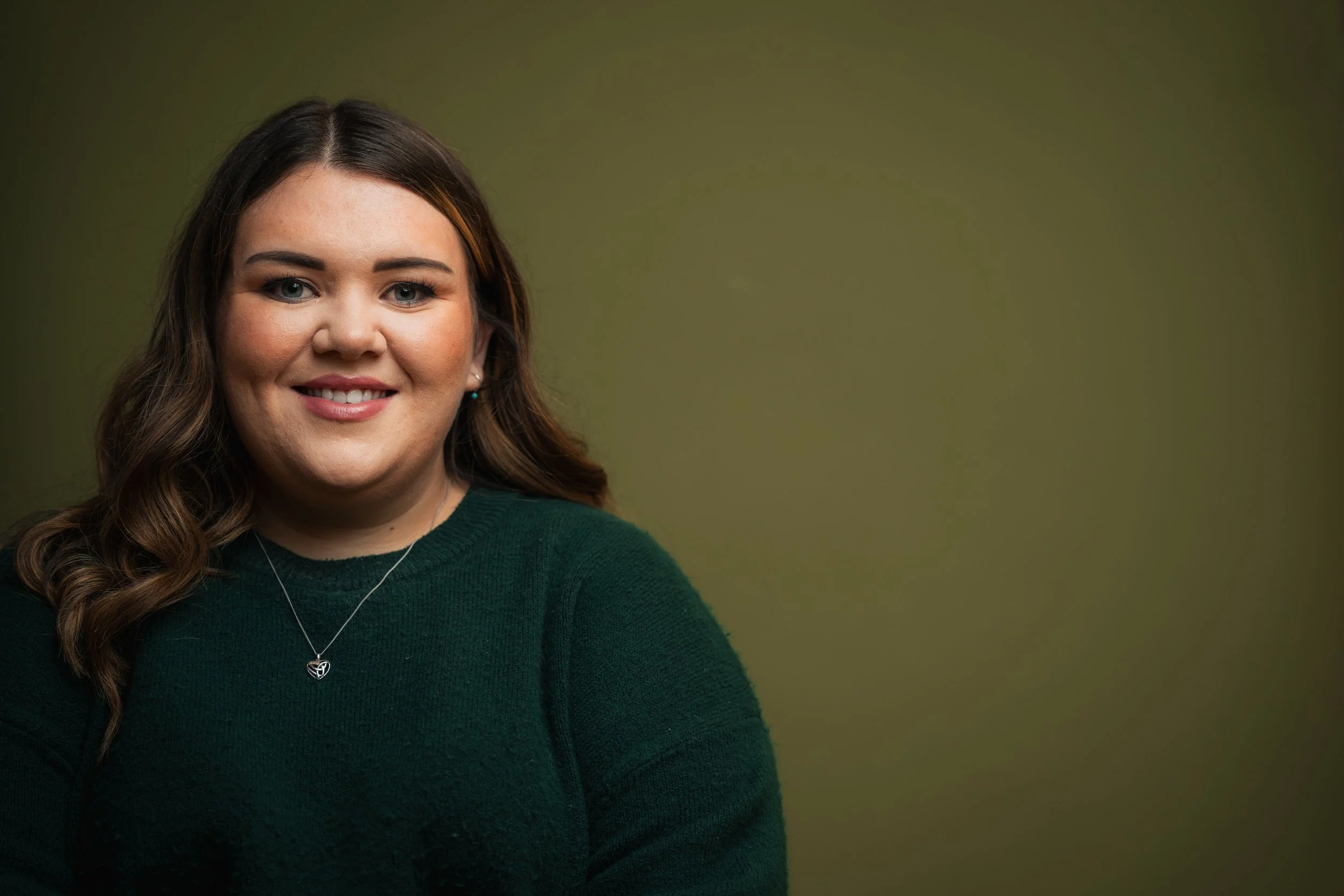 A young woman with long, wavy brown hair, wearing a dark green sweater and a silver necklace, smiling at the camera against a muted green background.