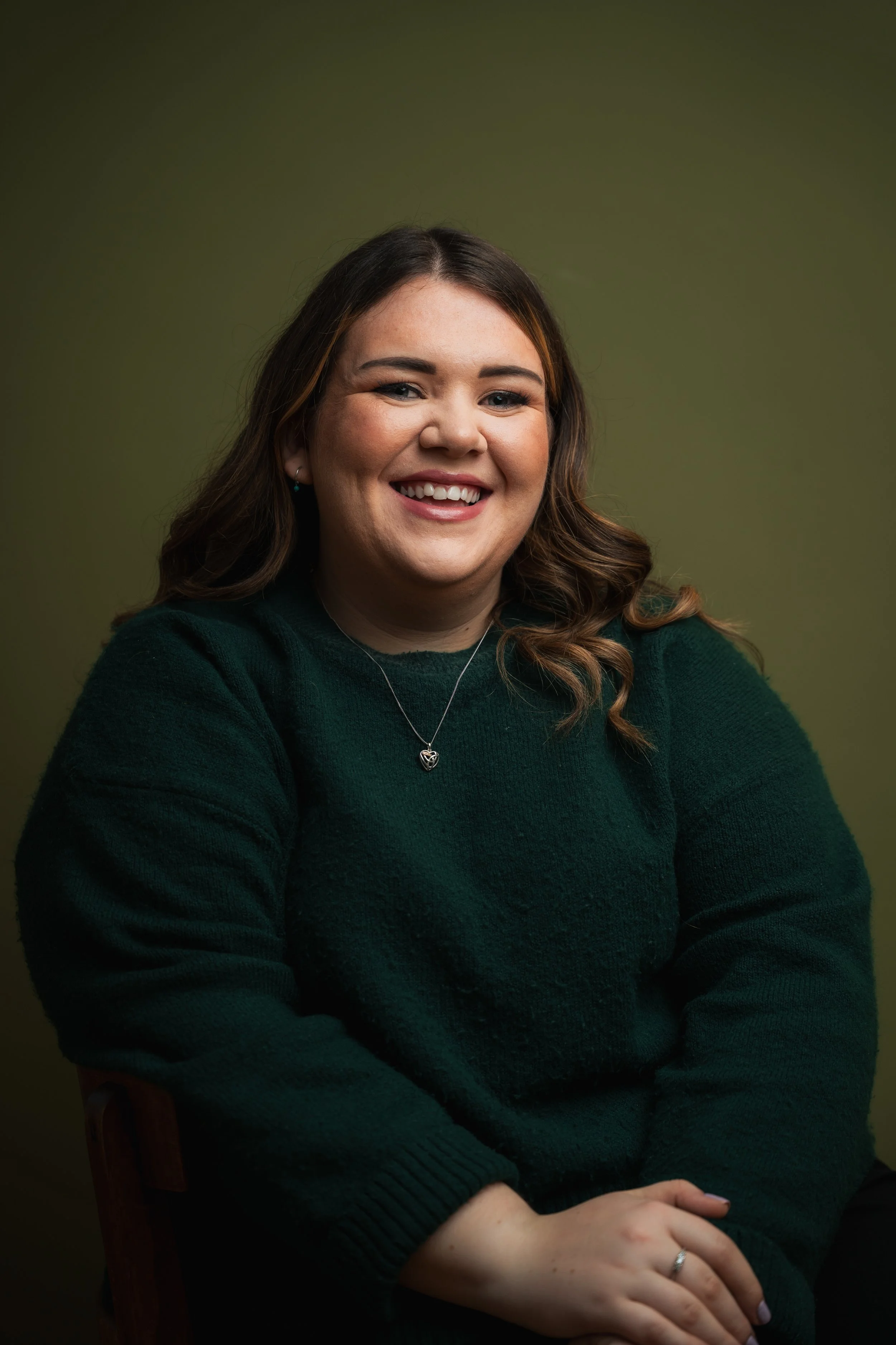 Portrait of a woman with brown hair smiling, wearing a dark green sweater and a silver necklace with a heart-shaped pendant, against a plain green background.