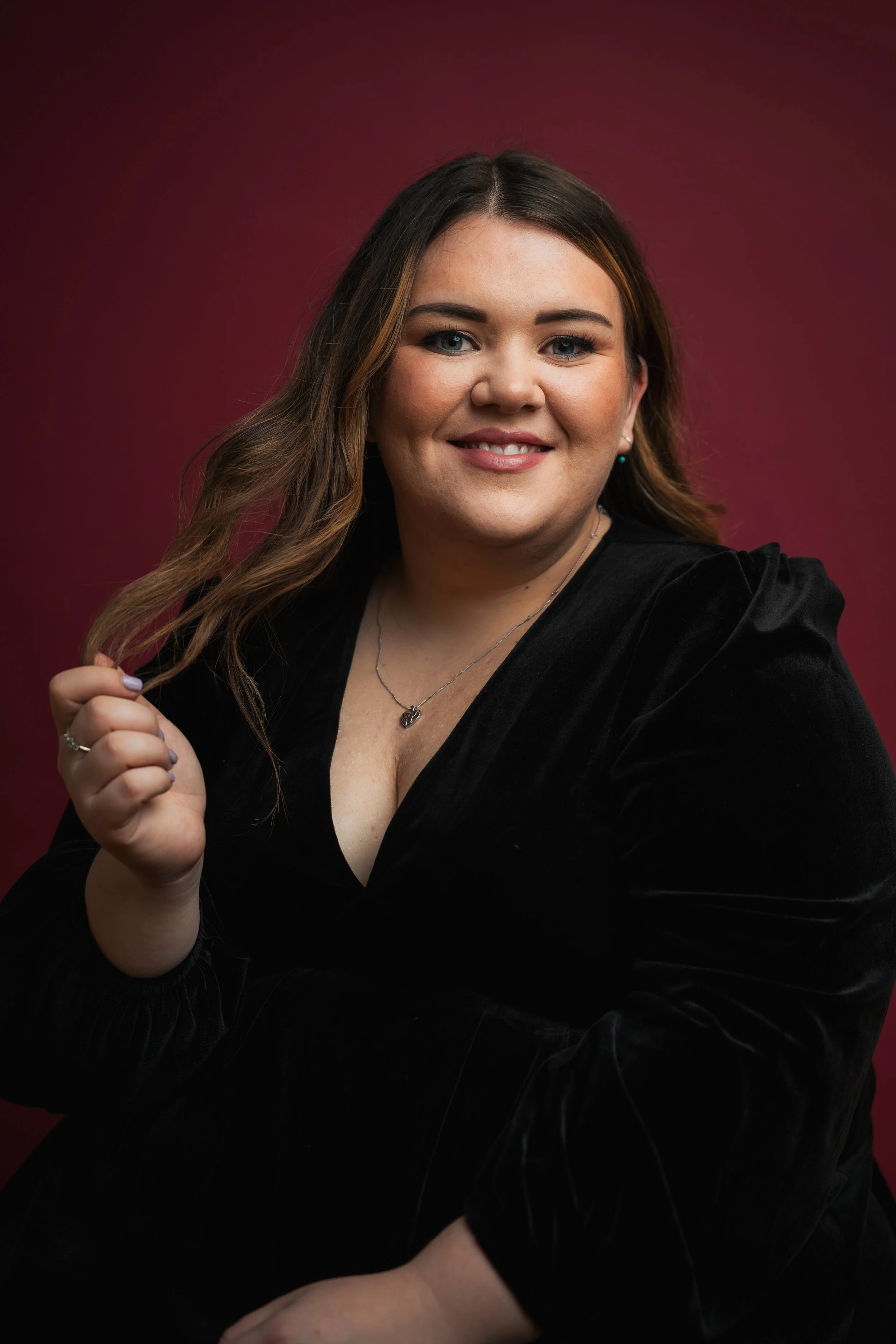 A woman with long brown hair and blue eyes wearing a black velvet dress, smiling, against a dark red background.