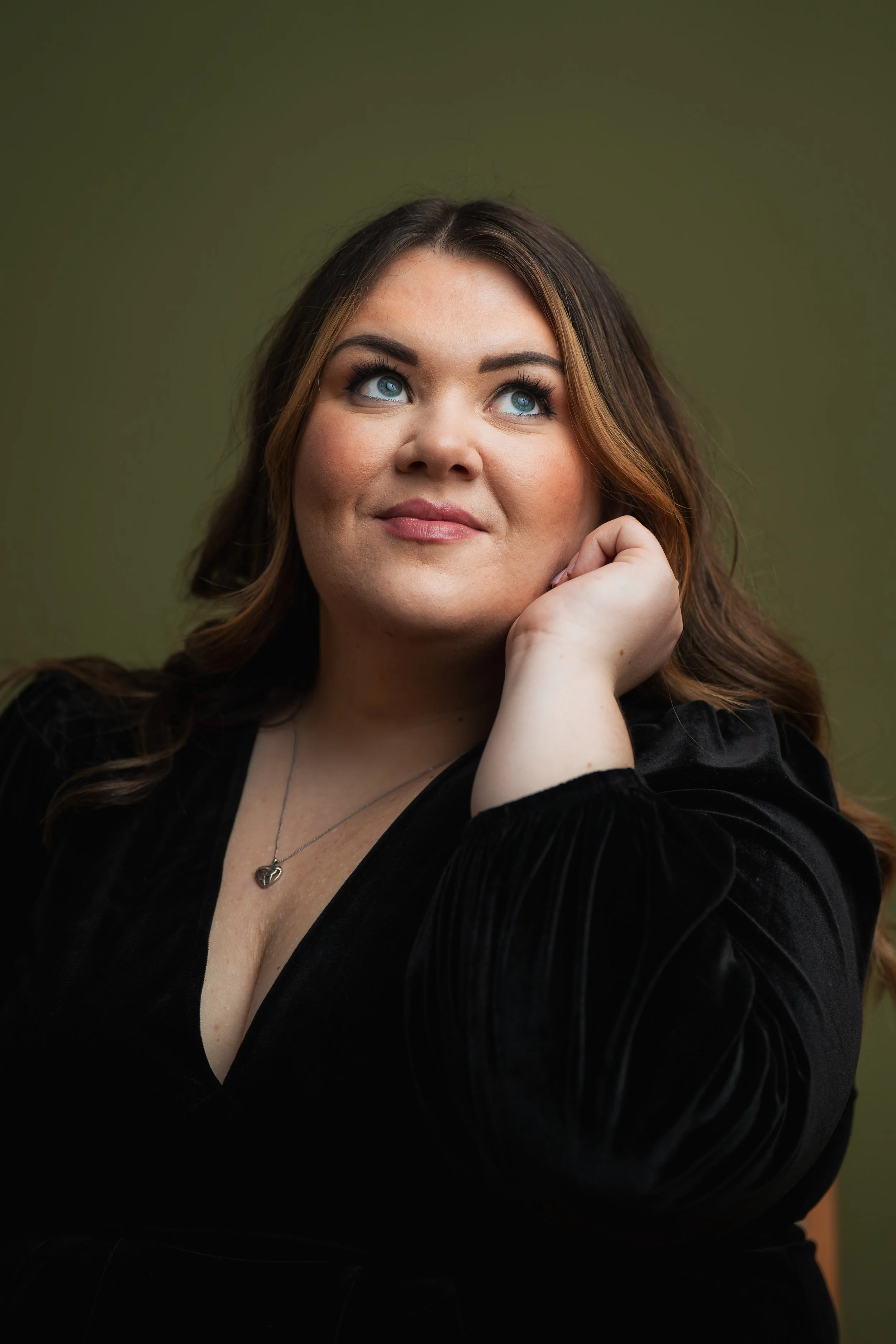 A woman with long brown hair and blue eyes, wearing a black top, resting her chin on her hand and looking slightly upward, with green background.