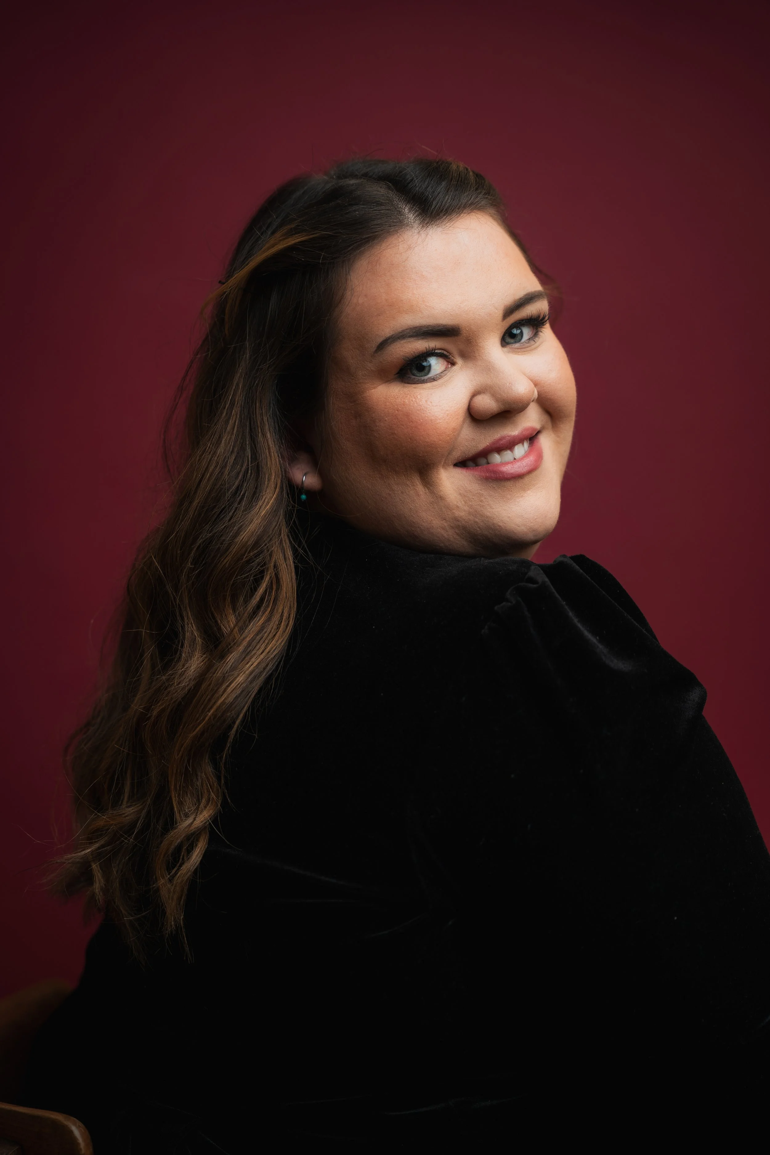 Portrait of a woman with long wavy brown hair smiling at the camera, wearing a black top against a pink background.