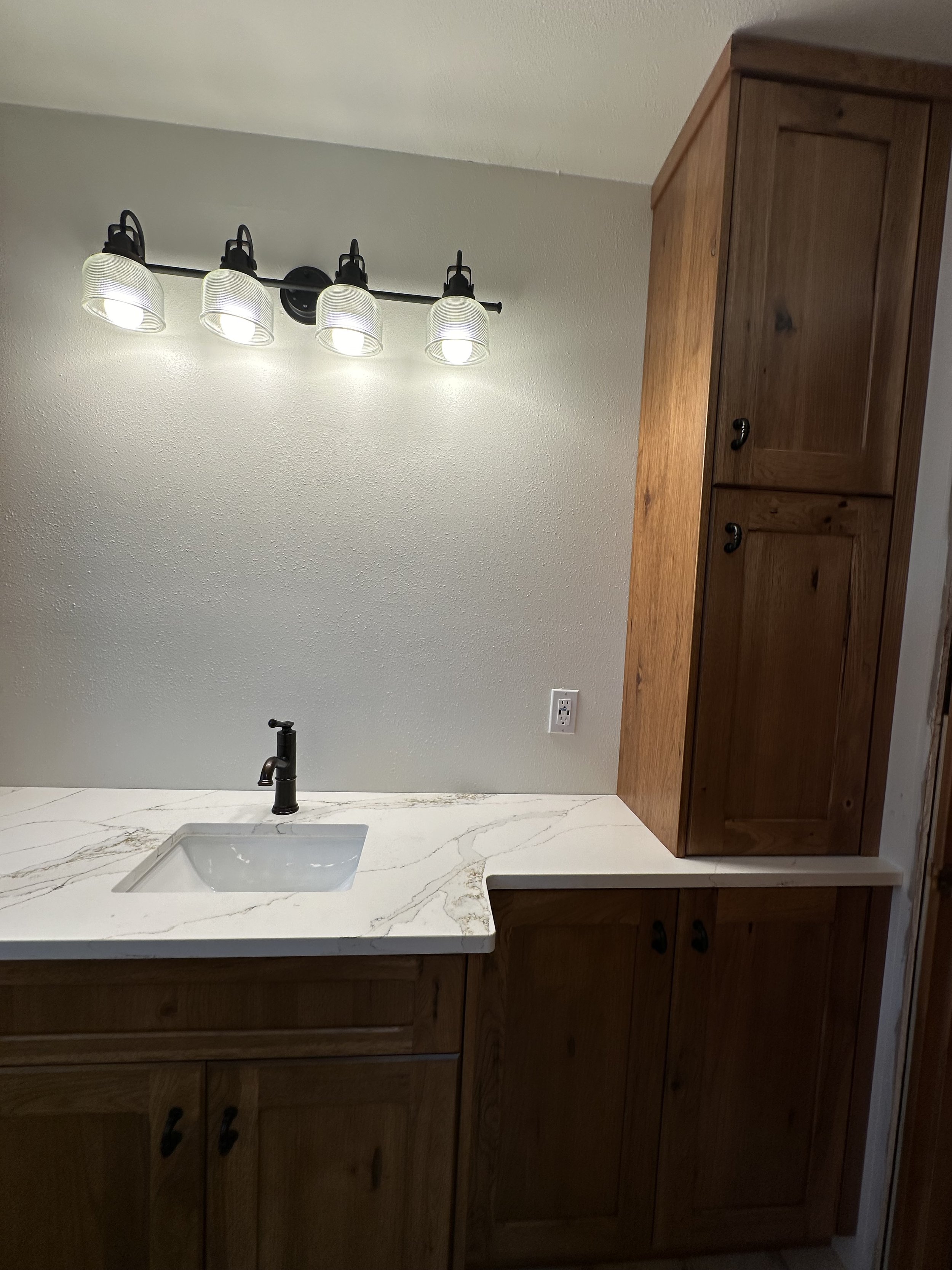 Bathroom vanity with white marble countertop, black faucet, wooden cabinets, and a wall-mounted light fixture with four glass shades.