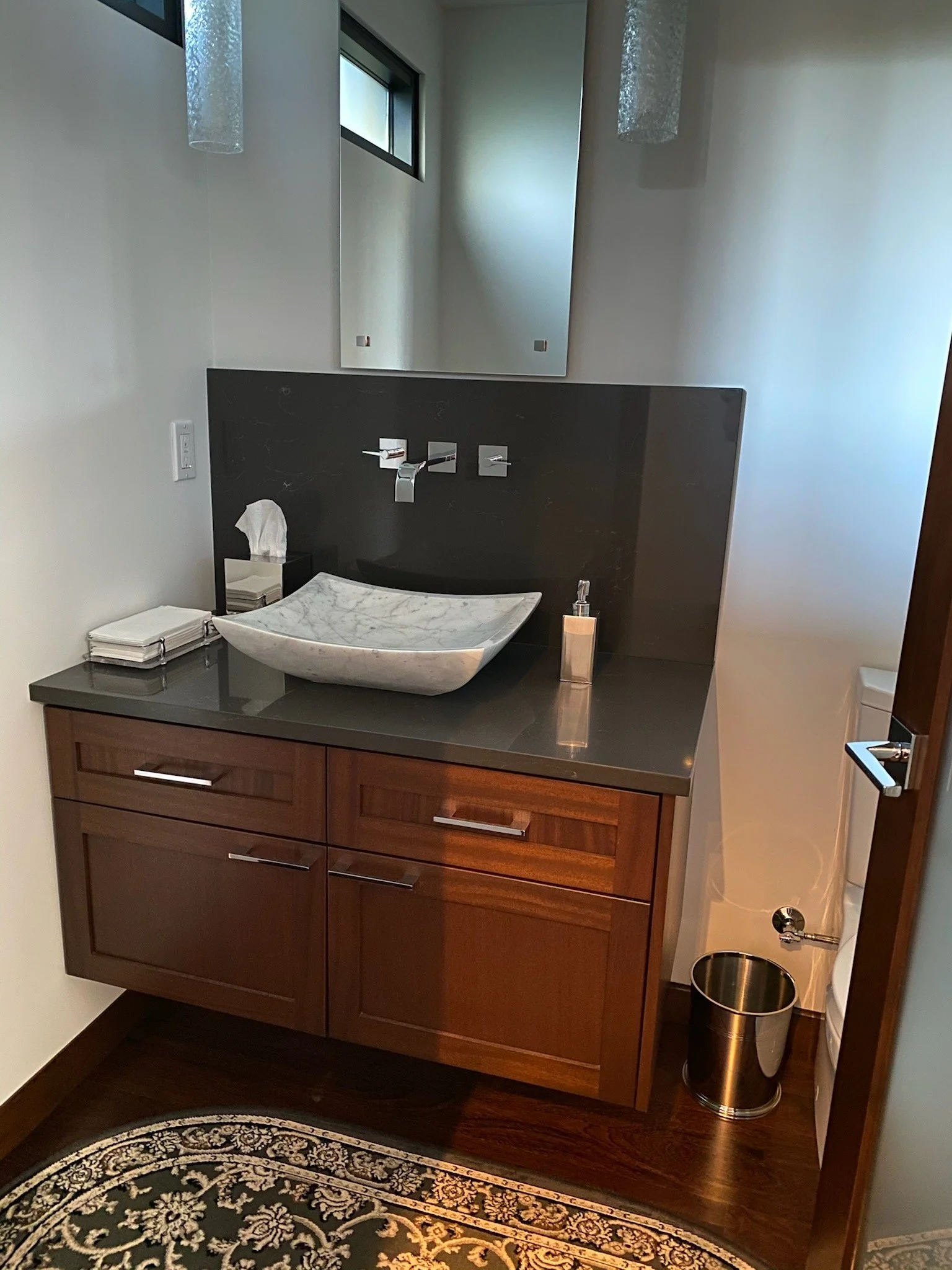 Bathroom vanity area with a wooden cabinet, a marble vessel sink, a wall-mounted faucet, a mirror, and some toiletries, with a small window and a decorative rug on a wooden floor.