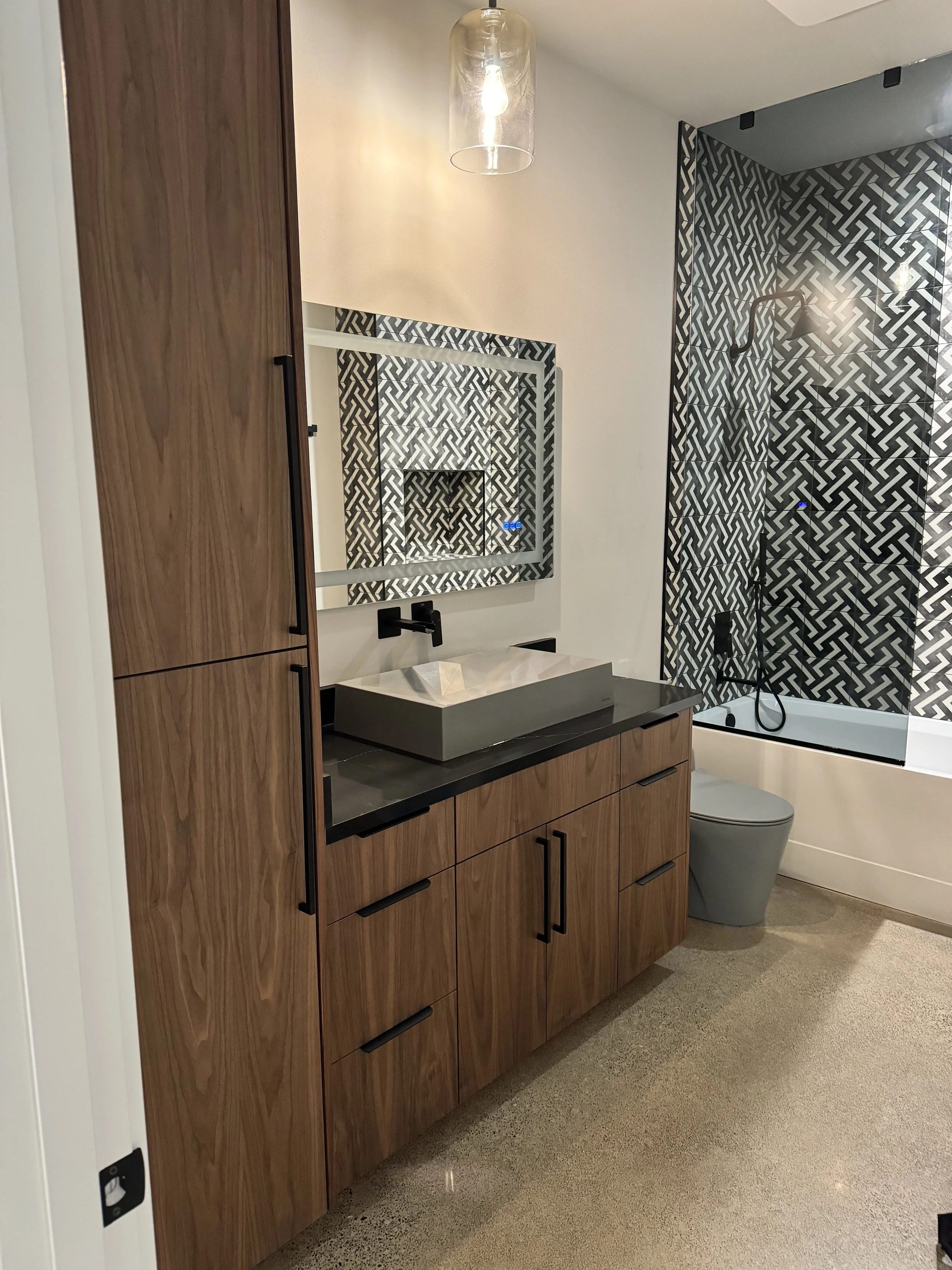 Modern bathroom with wooden cabinetry, a large mirror, a vessel sink, black fixtures, and a patterned black and white shower area with a glass door and separate toilet.