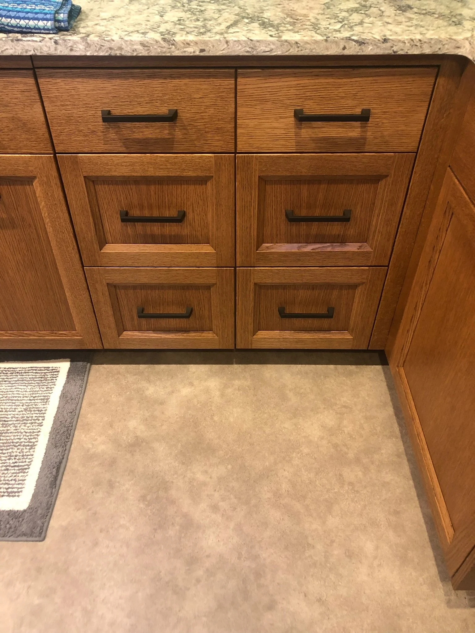 Wooden kitchen cabinets with black handles and a marble countertop, next to a carpeted area on a tiled floor.