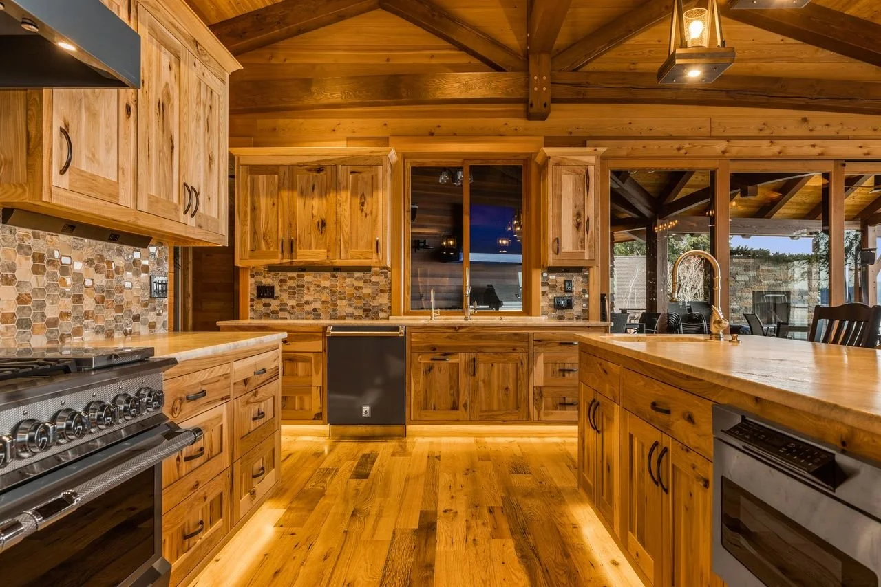 Rustic wooden kitchen with natural wood cabinets, a backsplash of small stone tiles, and modern appliances. Open layout with large windows and a view of an outdoor patio.