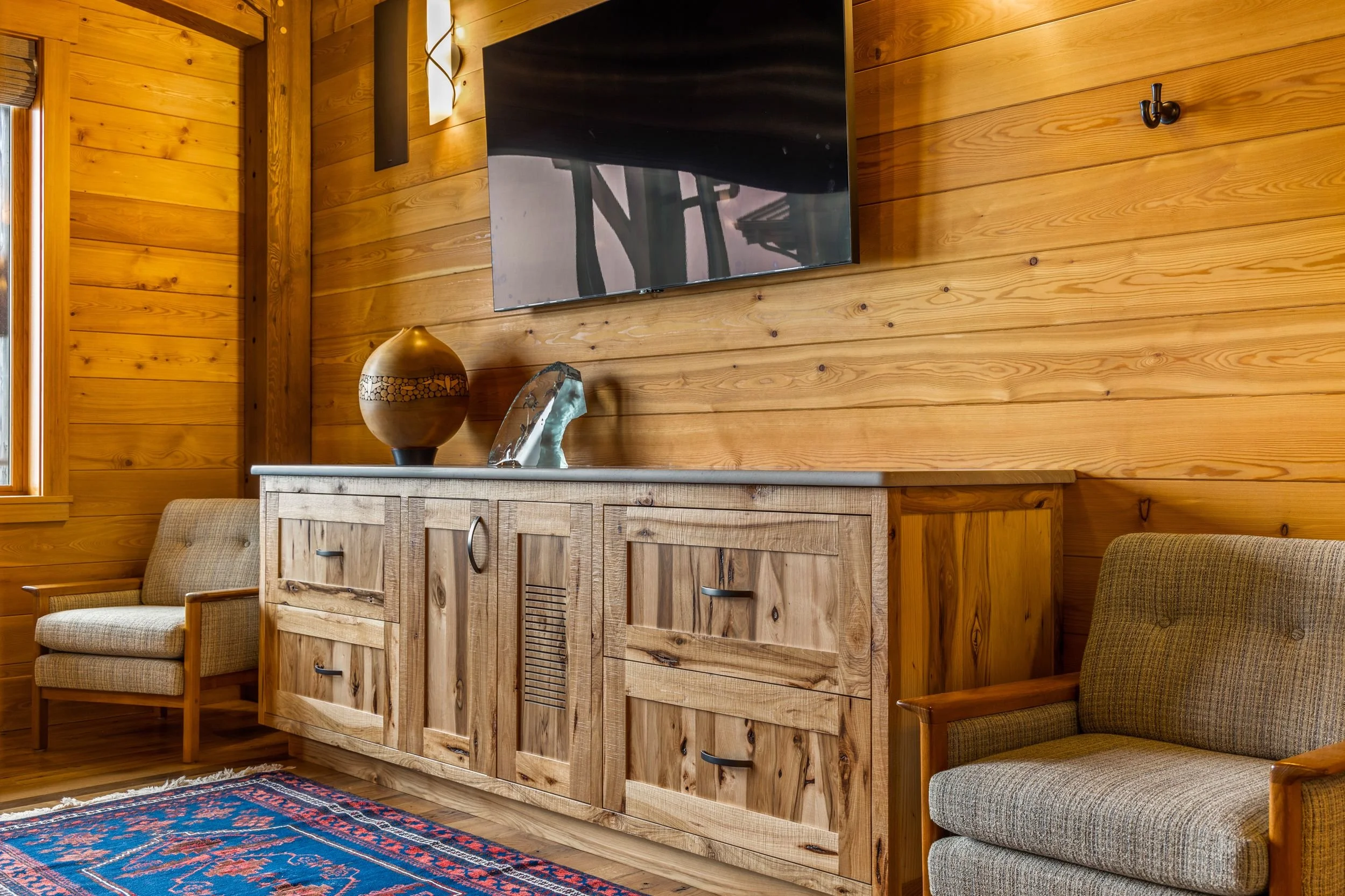 Living room with wooden walls, a wooden cabinet with decorative items, a wall-mounted TV, and two upholstered armchairs, one on each side of the cabinet, with a colorful rug on the wooden floor.