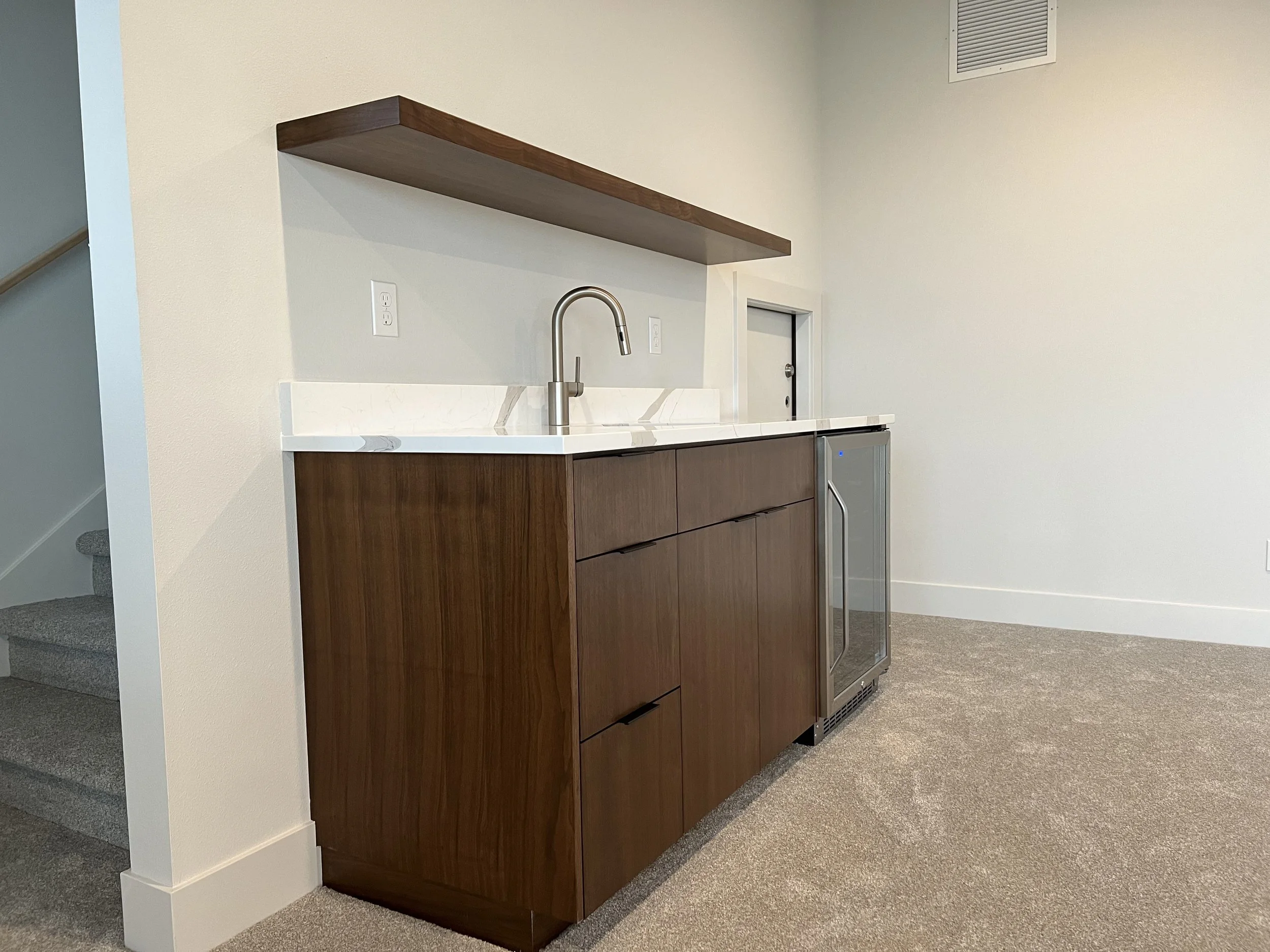 Kitchenette with wooden cabinet, Marble countertop, stainless steel sink and faucet, above shelf, electrical outlets, small refrigerator, beige carpeted flooring, white walls, partially visible stairs.