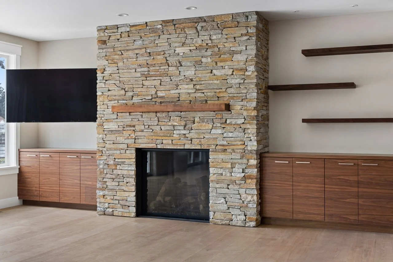 Interior view of a room with a stone fireplace, wooden shelves, and cabinets, and a large window allowing natural light.