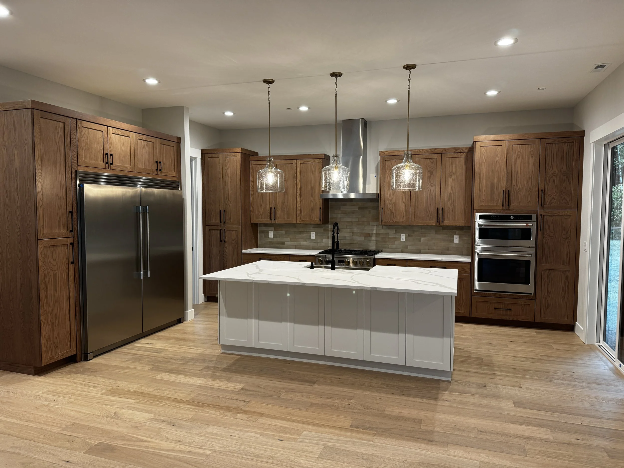Modern kitchen with wooden cabinets, stainless steel appliances, a white island with marble countertop, three hanging pendant lights, hardwood floors, and a sliding glass door.