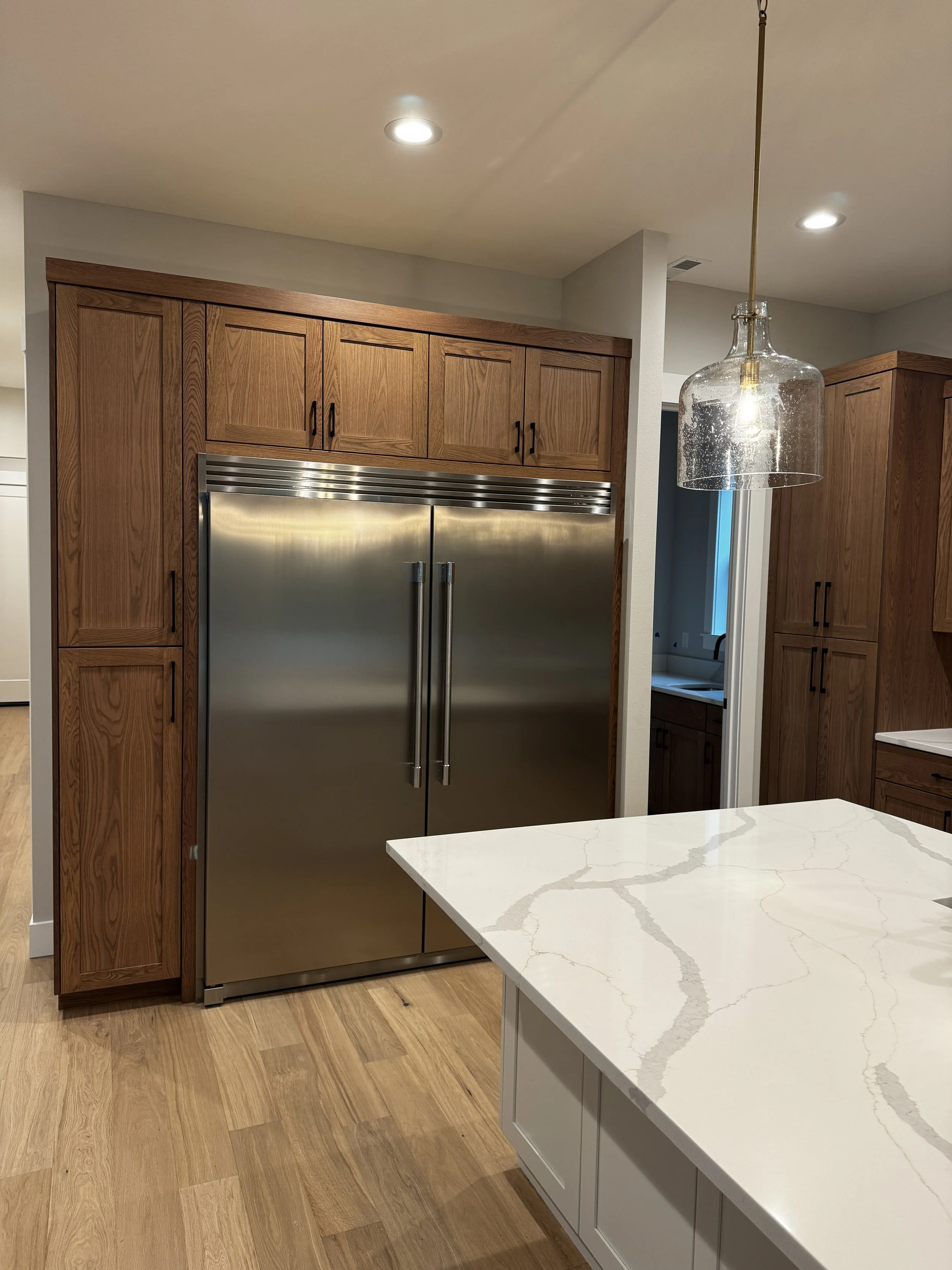 Stainless steel double-door refrigerator surrounded by wooden cabinets in a modern kitchen, with a white marble countertop with gray veining and a hanging glass pendant light.