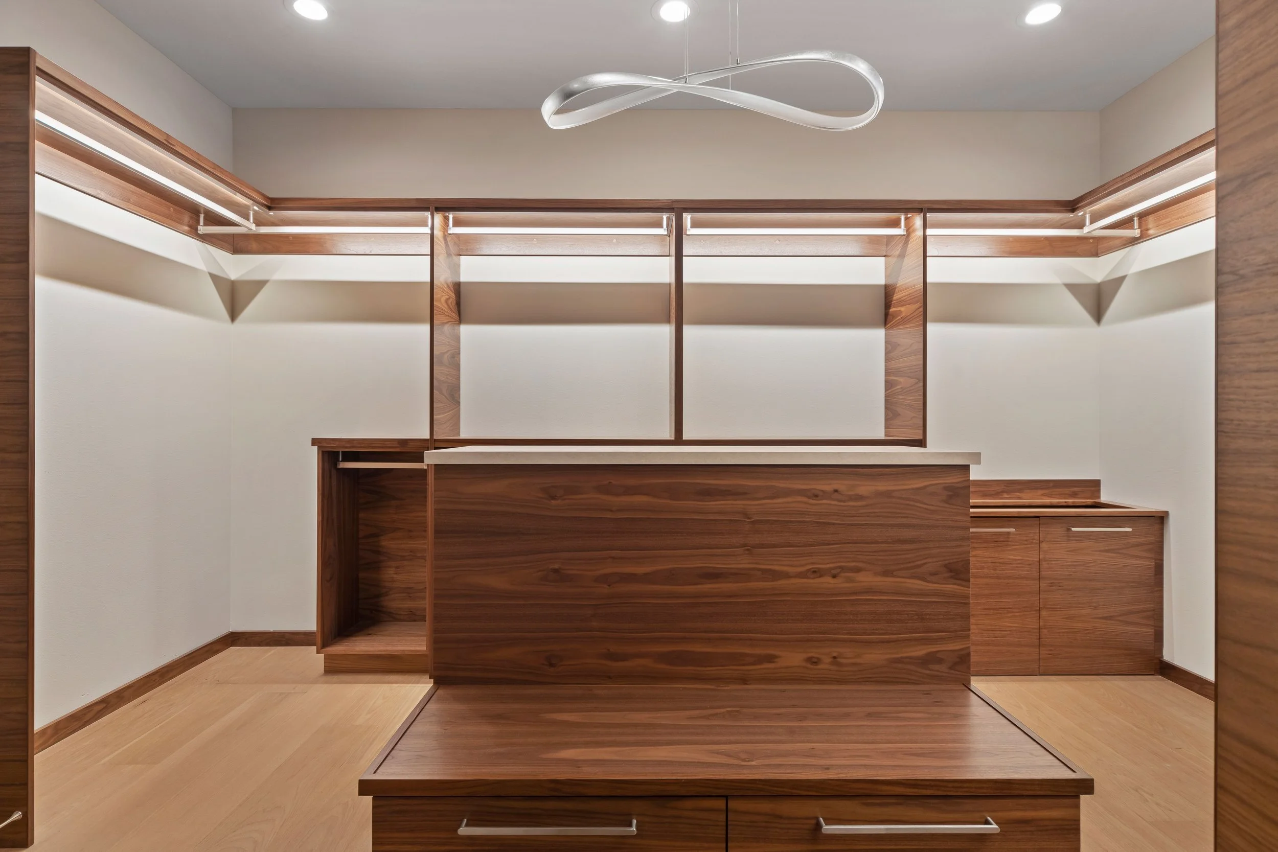 Empty walk-in closet with wooden shelves, drawers, and a hanging rod, illuminated by ceiling lights.