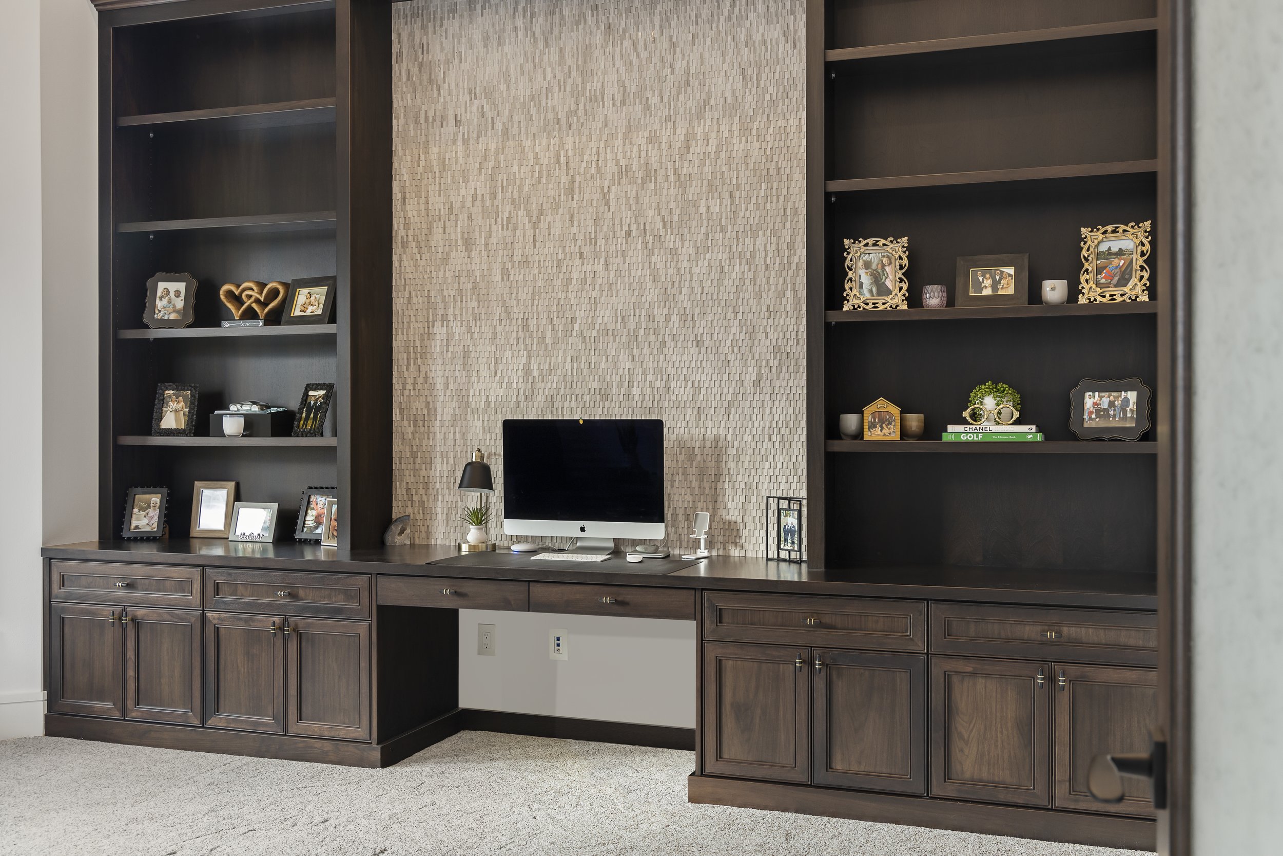 Home office with dark wood built-in shelves and cabinets, a desktop computer, desktop lamp, and various framed photographs and decorative items on the shelves. The background features a textured beige wall.