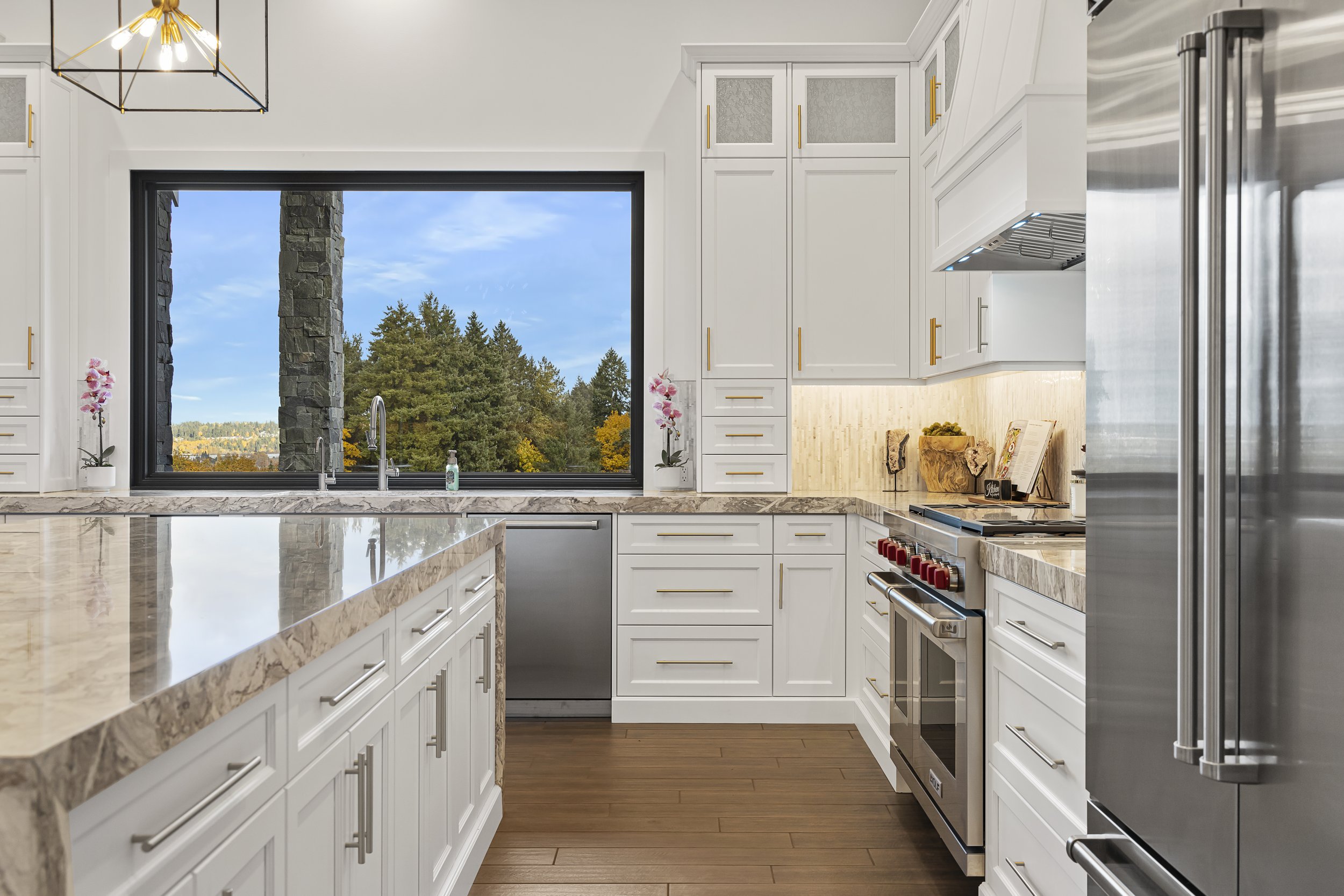 Modern kitchen with white cabinetry, marble countertops, stainless steel appliances, and a large window overlooking trees.