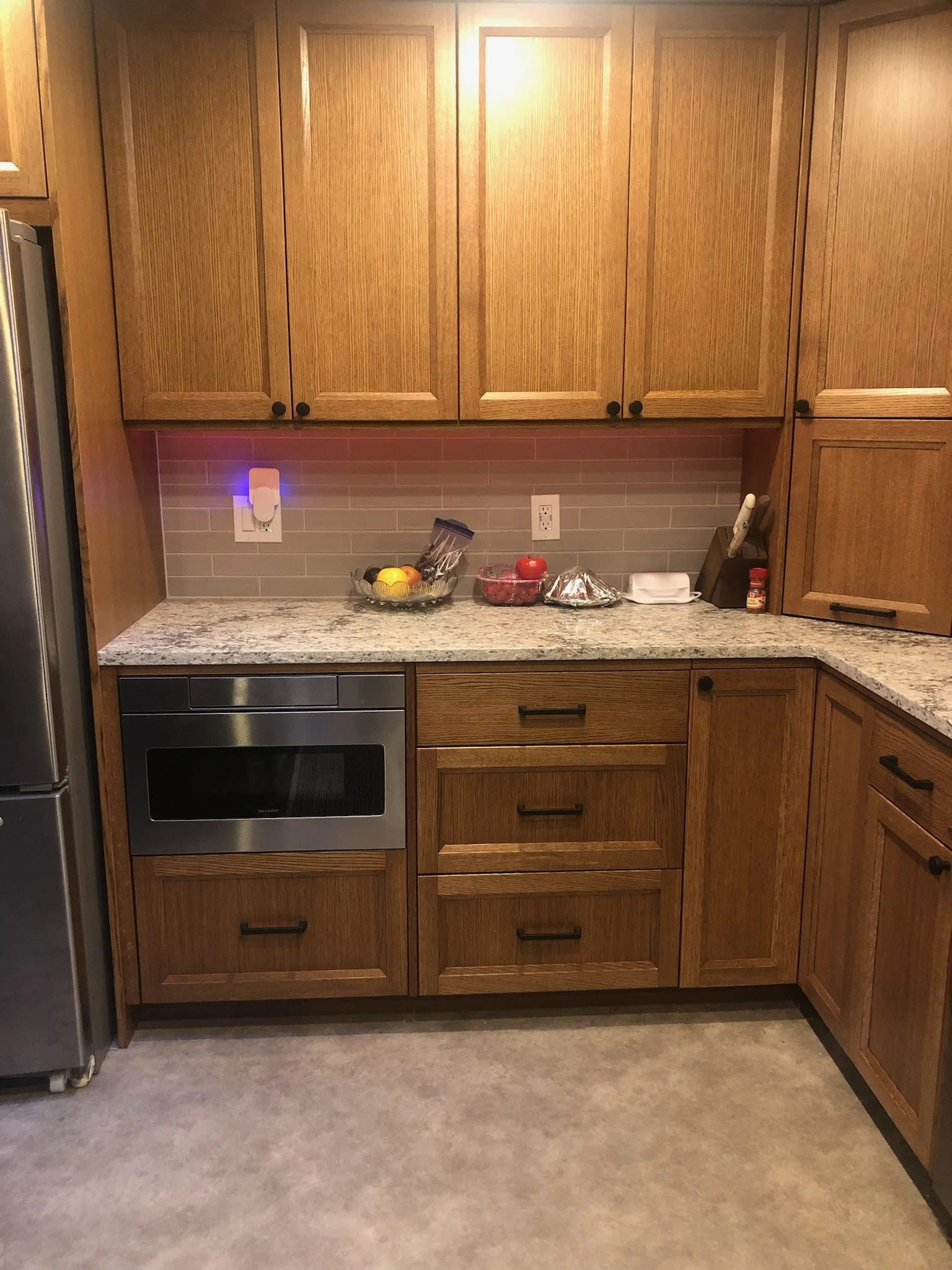 Wooden kitchen cabinets above granite countertop with various kitchen items, including a fruit bowl, apples, foil-wrapped items, plastic containers, knives, and spices.