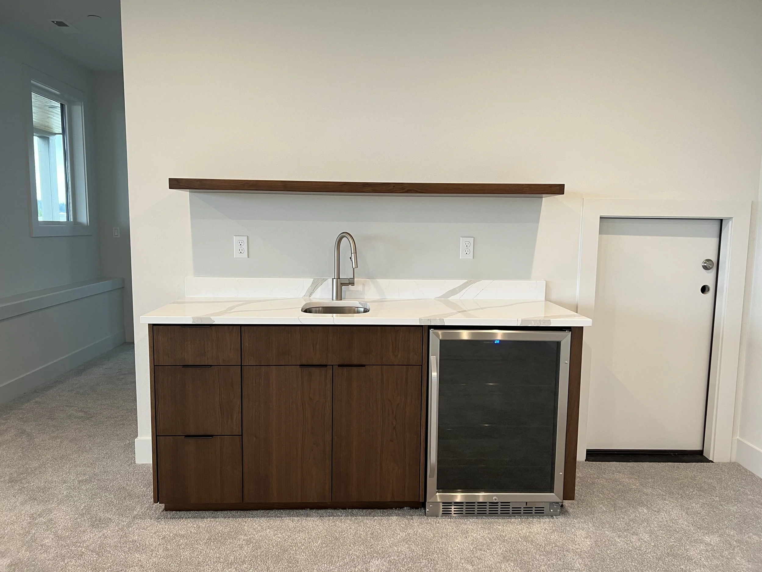 A small kitchenette with wooden cabinets, a white marble countertop, a stainless steel sink with a modern faucet, a built-in mini fridge with a dark door, and a wooden floating shelf above. There are two electrical outlets on the wall behind the coun