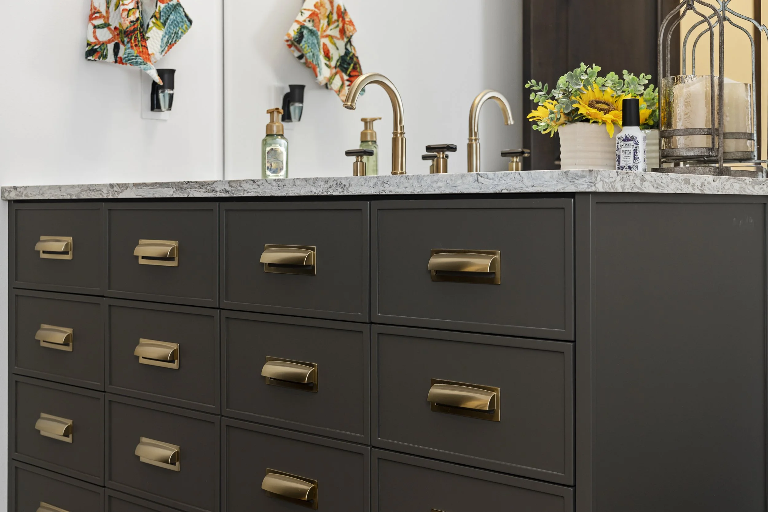 A kitchen vanity with grey drawers and gold handles, topped with a marble countertop. There are two sinks with gold faucets, and various decor items including a sunflower plant, a bottle of soap, a lantern, and small towels hanging on the wall.