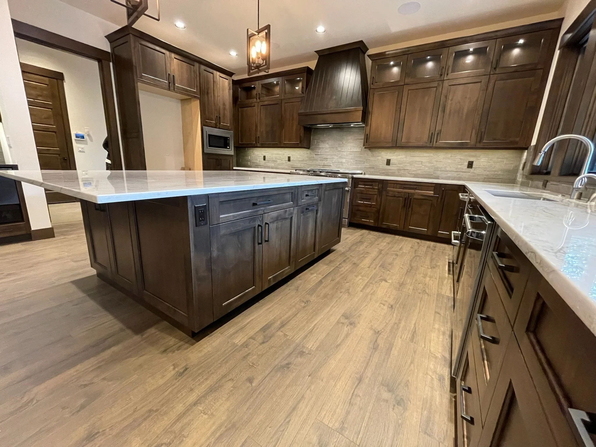A spacious kitchen with wooden cabinets, a marble island in the center, and hardwood flooring. The room features a range hood, microwave, and modern fixtures, with recessed lighting and pendant lights overhead.