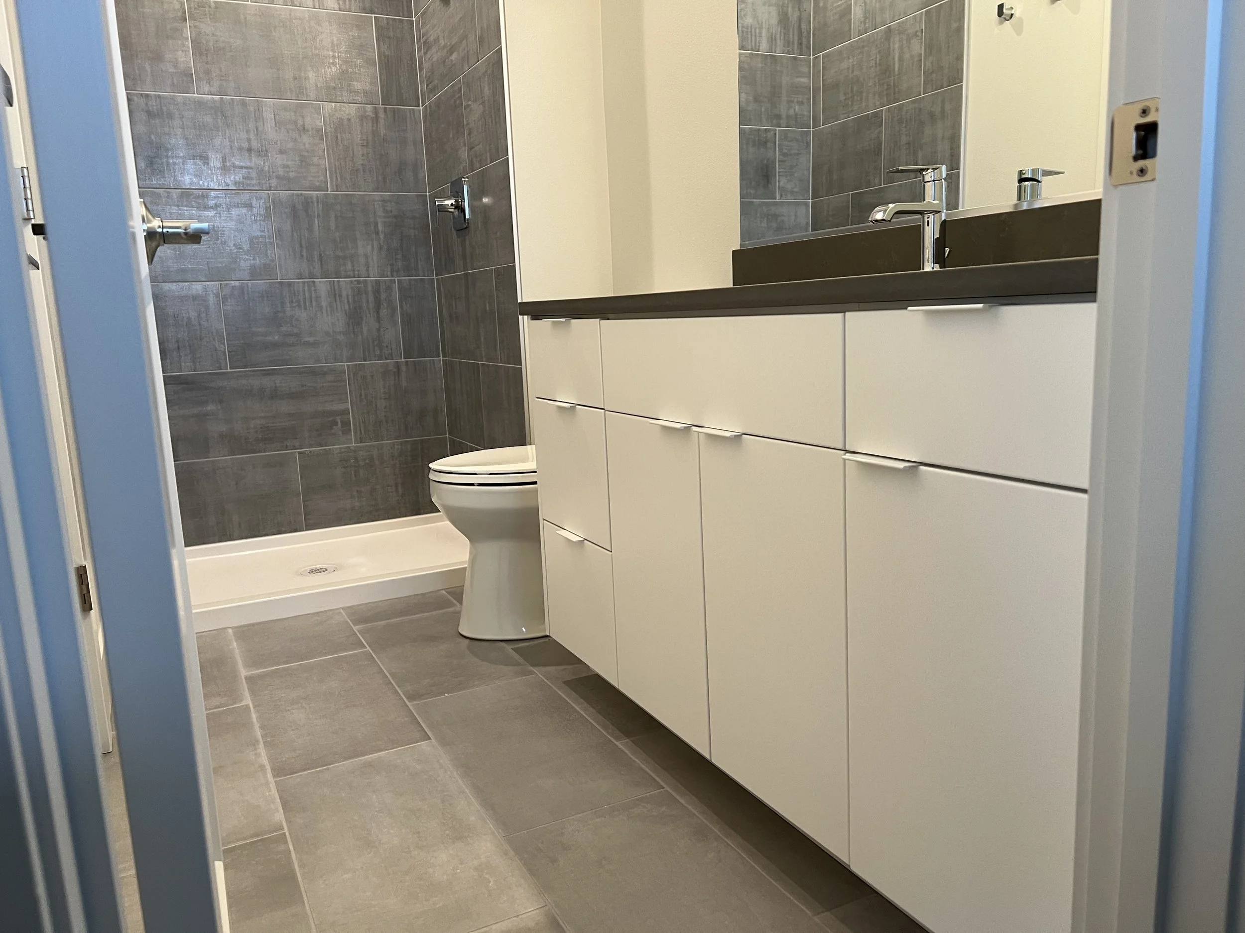 Modern bathroom with gray tiled shower area, white toilet, and a white vanity with a dark countertop and two sinks.